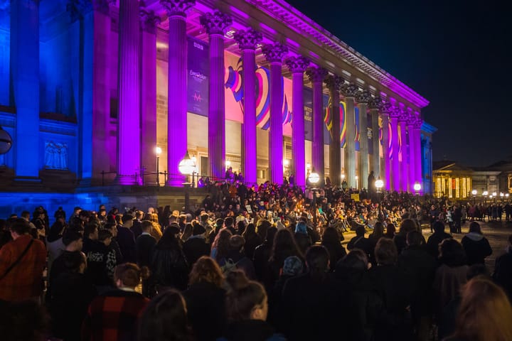 St George's Hall lit up in the blue and pink colours of the trans flag. A large crowd has gathered on the steps with signs an