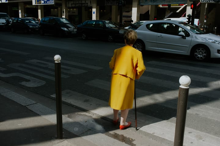 A woman in a yellow outfit and red shoes crosses a road.