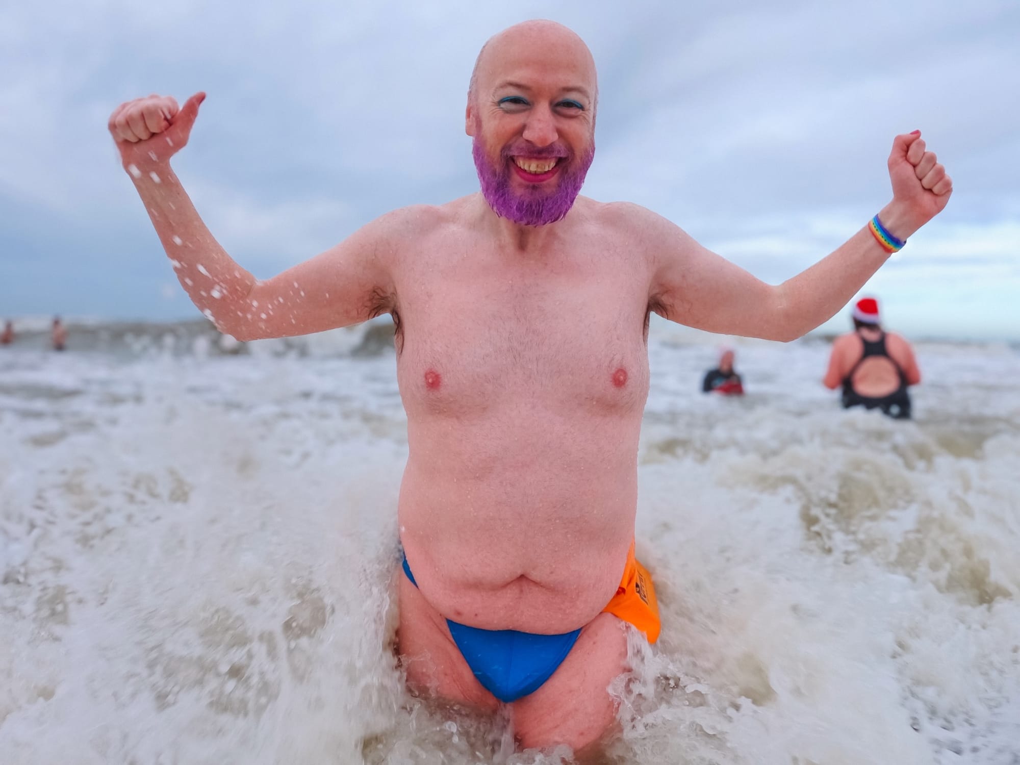 A happy non-binary person wearing only a tiny blue thong raises their fists in the air while the waves crash around them.