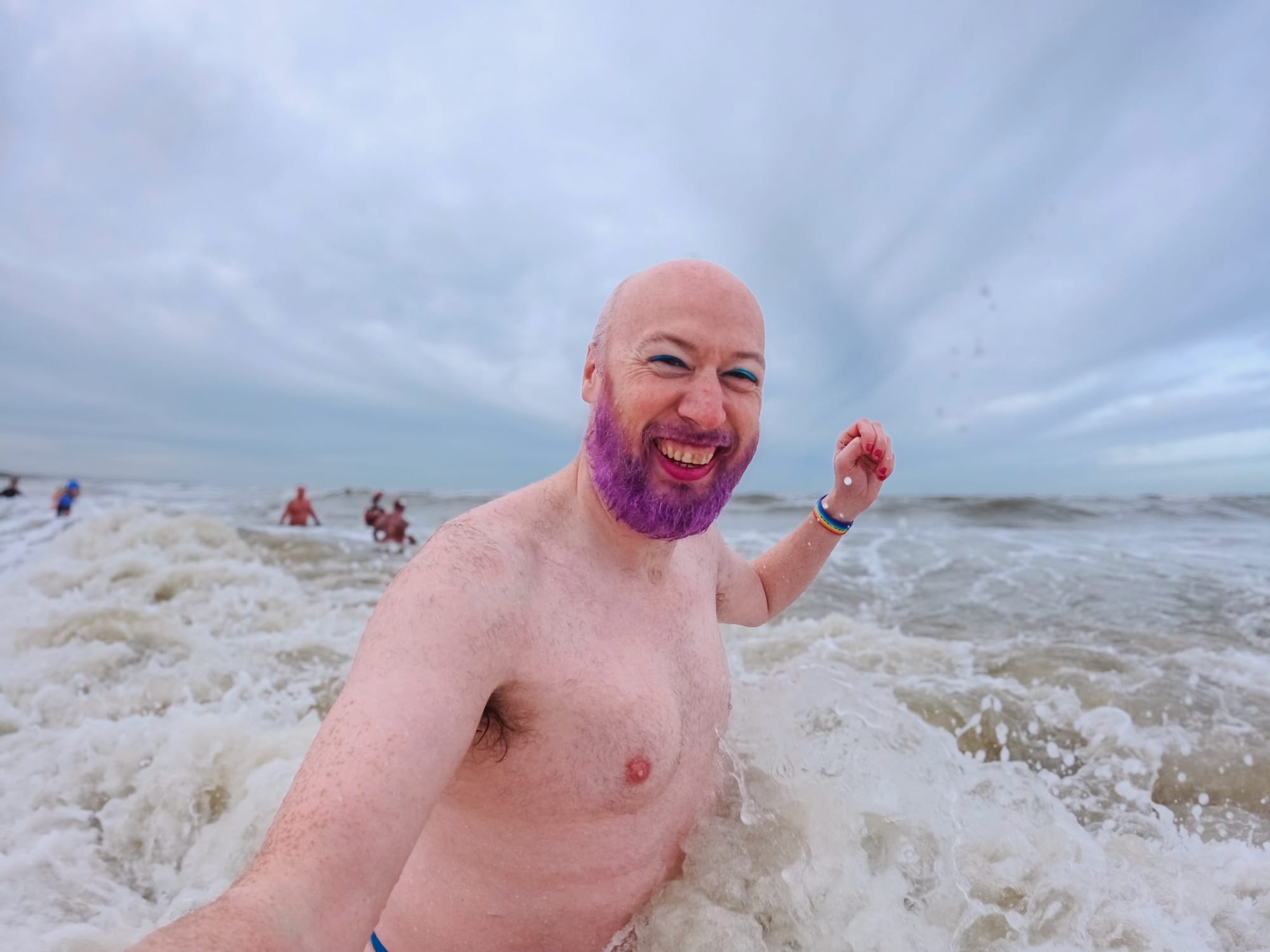 A happy non-binary person laughs as a wave crashes into them. They have a purple beard and blue eye liner on.