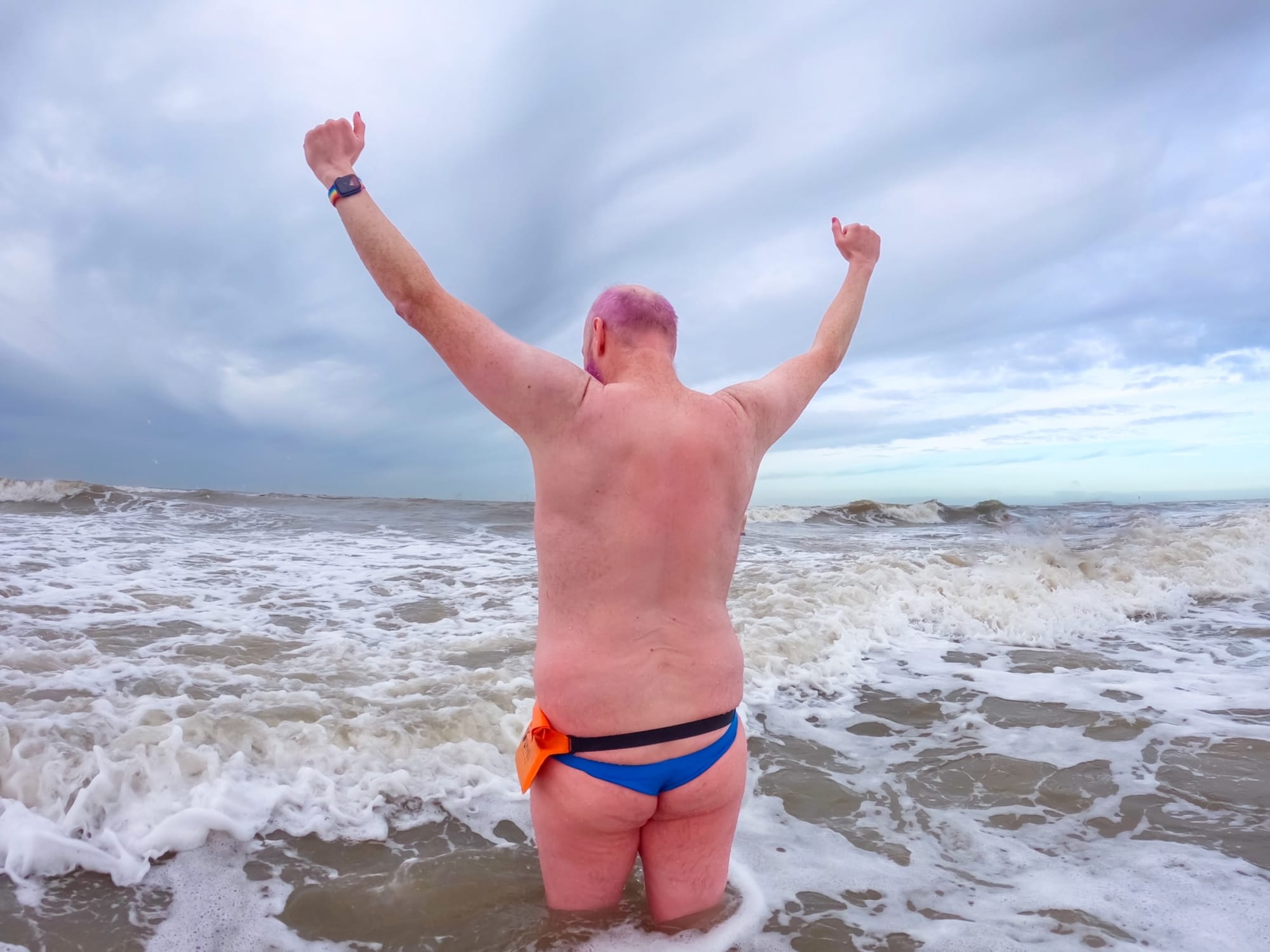 A male bodied non-binary person standing knee deep in the water. They have their back to the camera and are wearing only a blue thong.