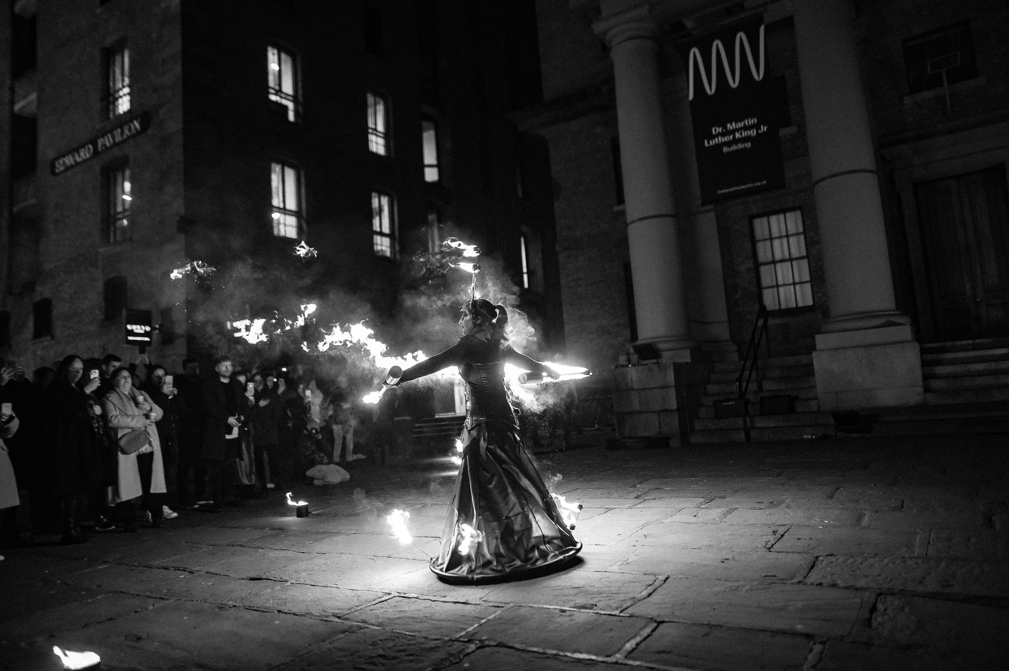 A woman wears a dress with fire surrounding her feat and sticks of flames on her headdress. She has firey poles that spew fire as she waves them.