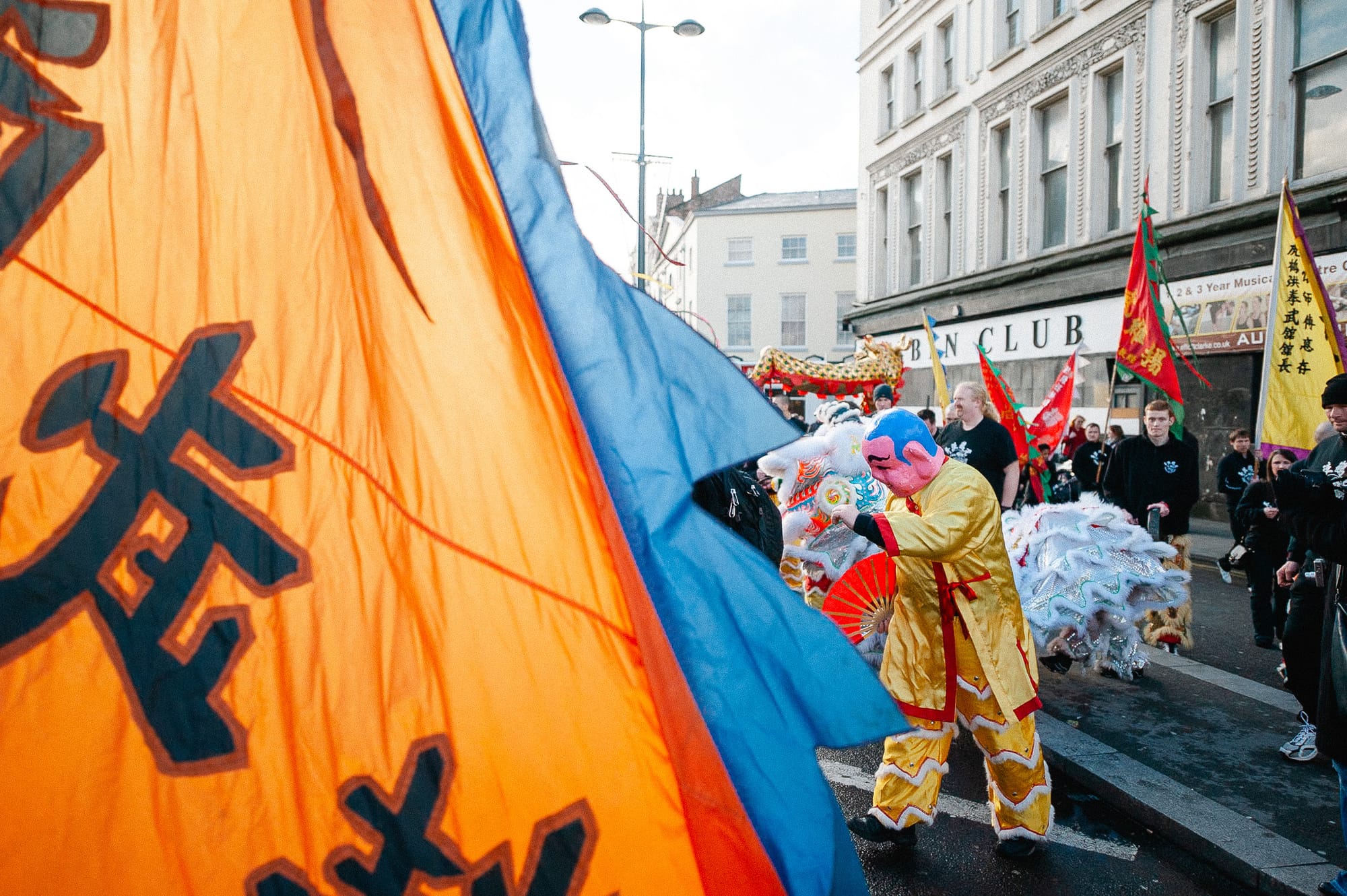 A person in a Buddha mask stands in the distance while flags fly close up to the lens of the camera.