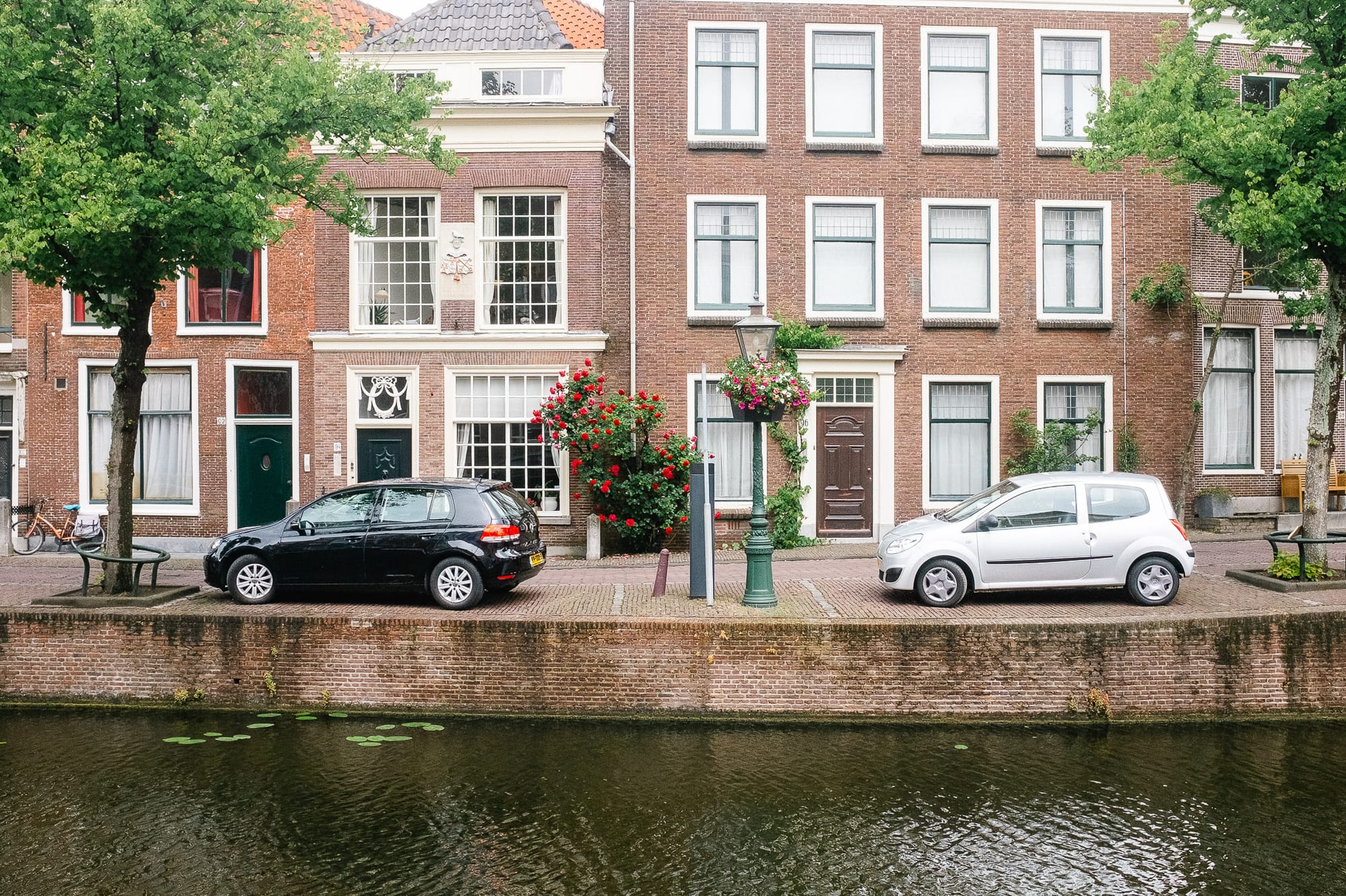 Standing on the other side of a canal in Leiden, Netherlands. There are cars parked next to the canal and lots of greenery. Big trees and bushes by the houses.