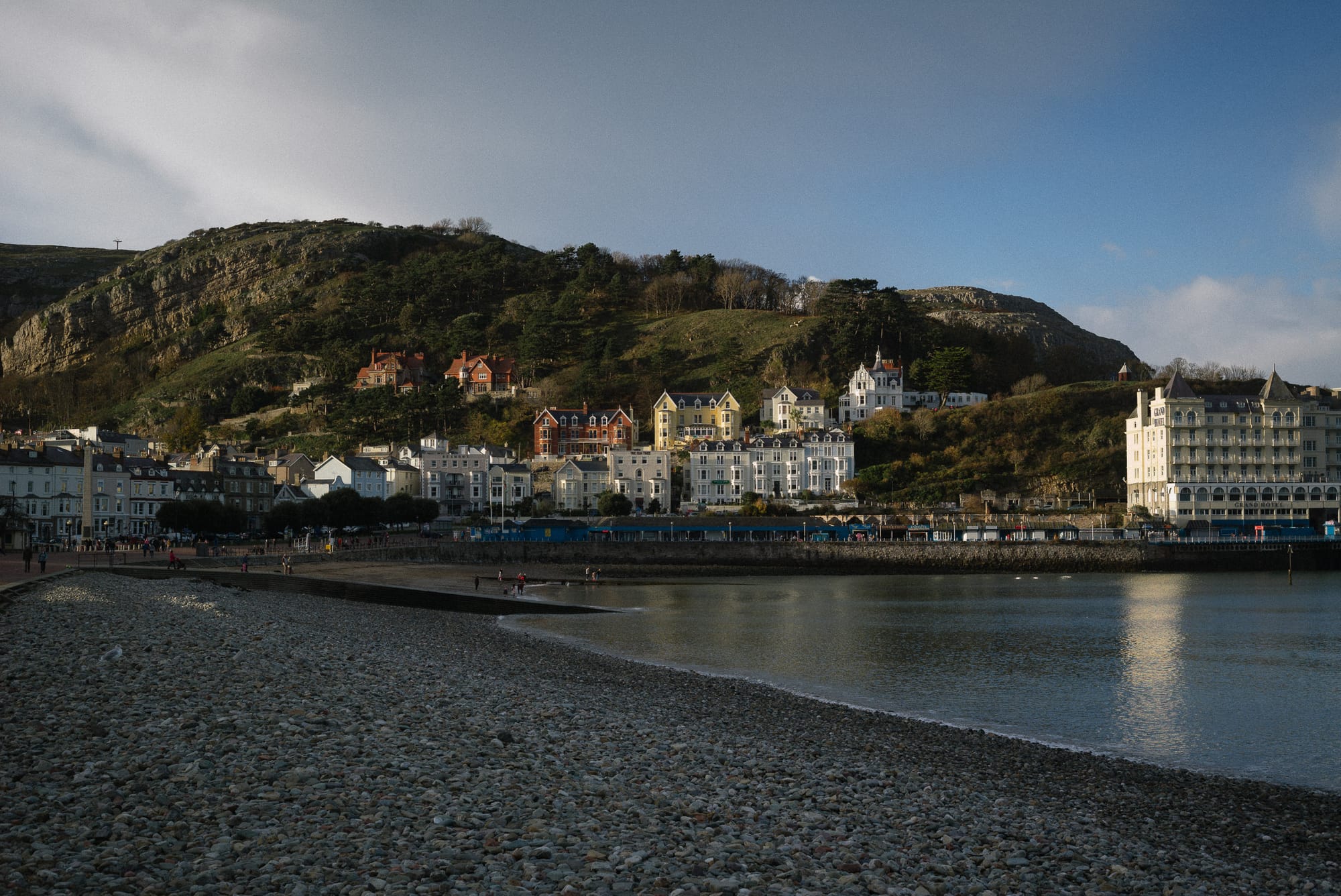 The tide calmly rolls in onto a pebble beach. In the background there is a large hill with white houses at the bottom and a large hotel.