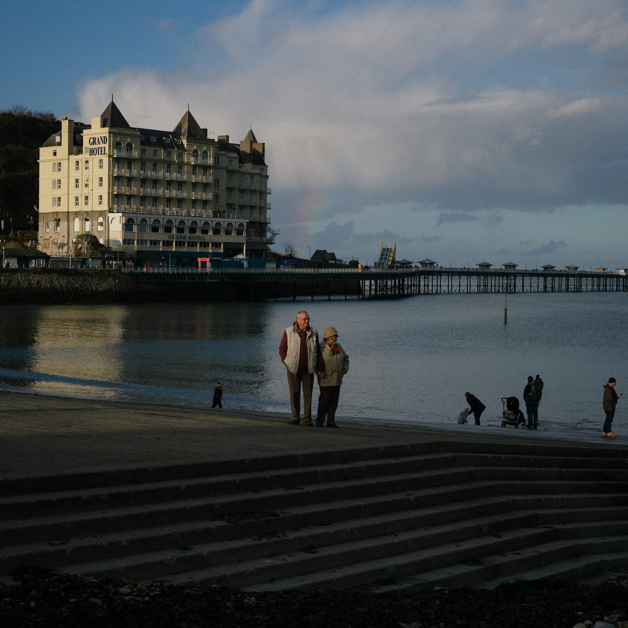 Two older people stand by steps on a beach in a spot of sunlight at the waters edge. In the background there is a large hotel and pier.
