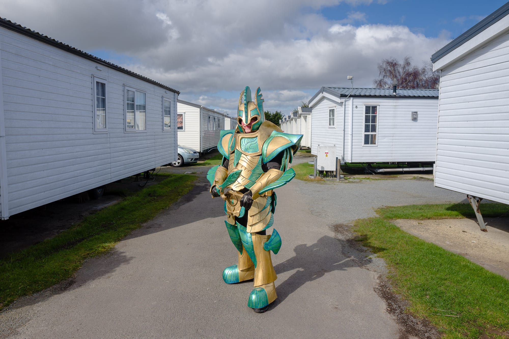 A man wears a green and gold coloured epic suit of armour in a caravan park.