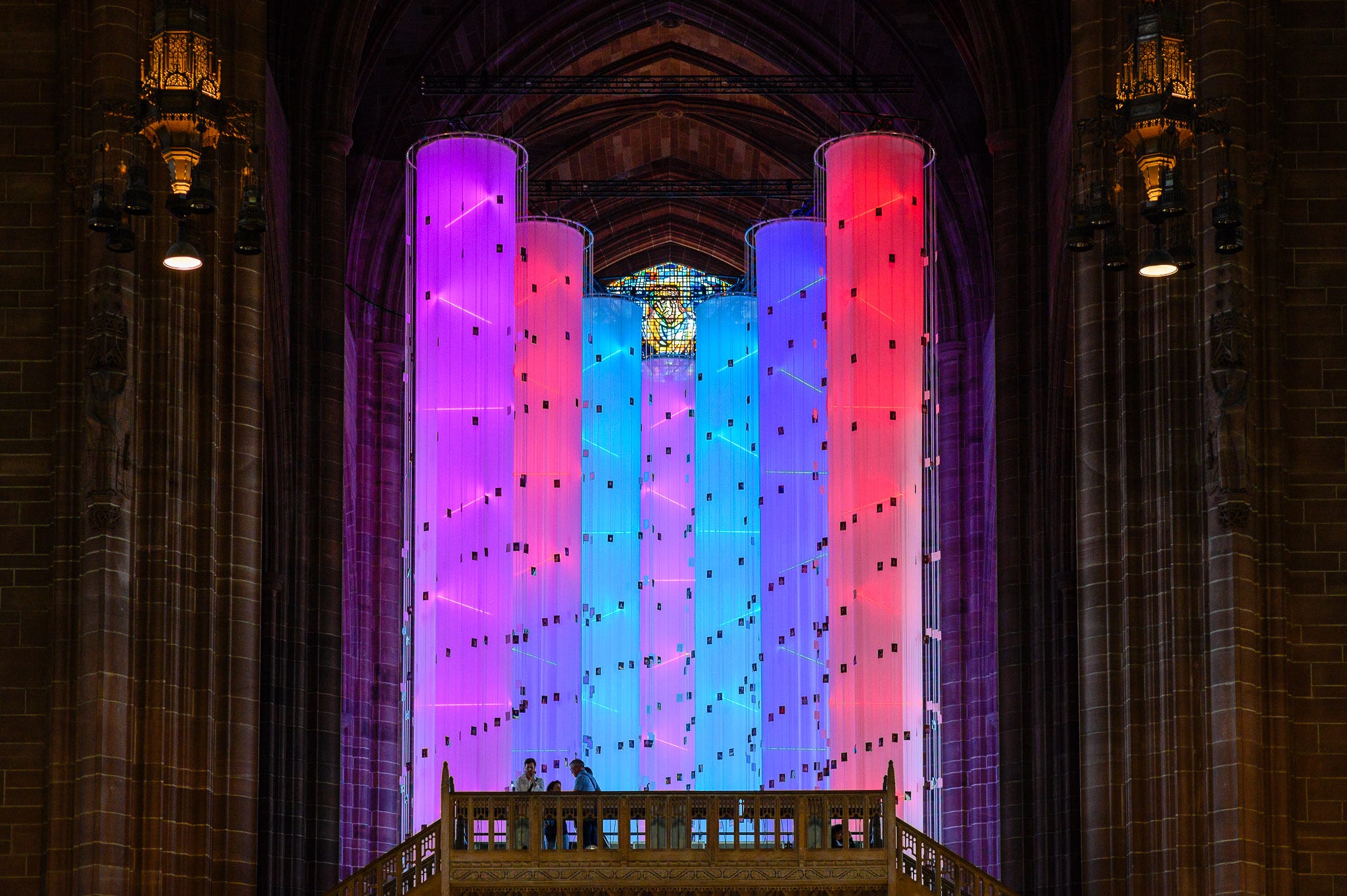 Standing beneath a bridge in the cathedral there are people crossing it to get a good view of the rainbow columns. 