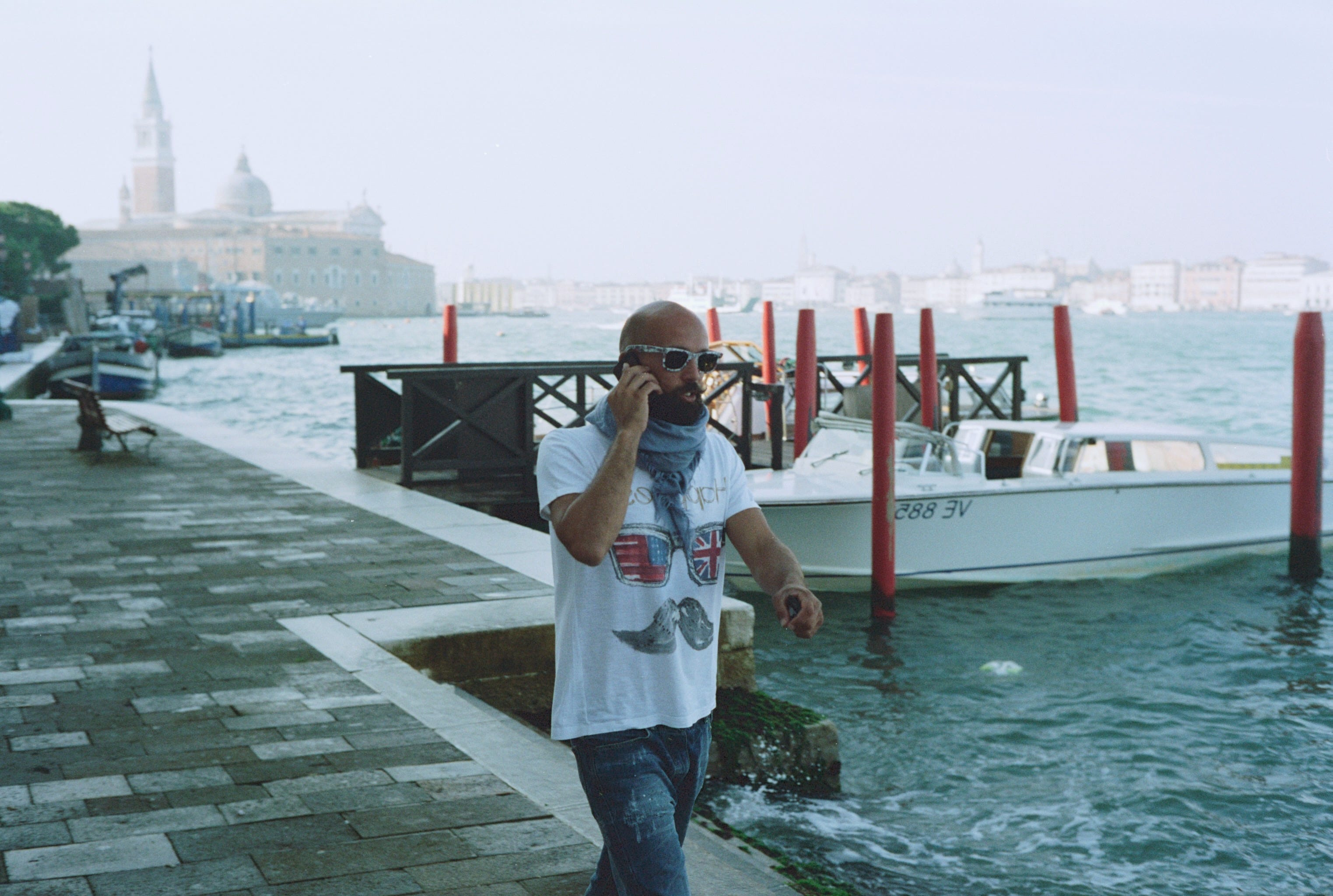 A bald man with a beard talks on his phone as he passes water taxis in Venice.