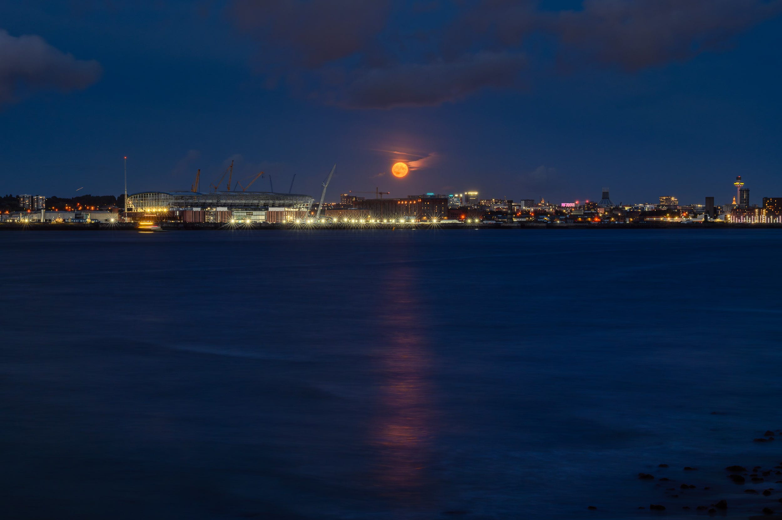 Super blue moon rises through the clouds over the Liverpool waterfront. The river is calm and the reflection of the orange moon stretches across it.