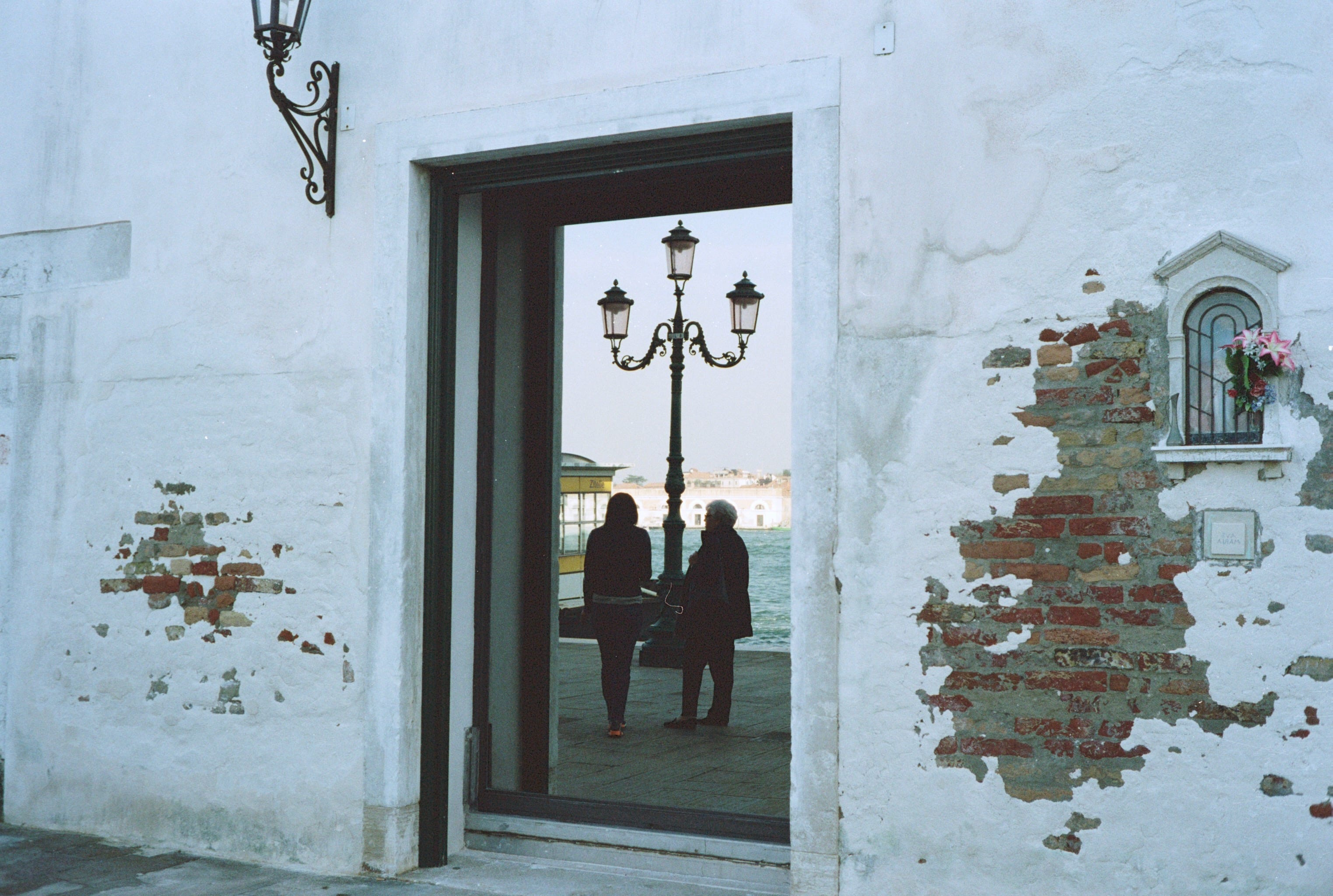 At the side of a building, people are reflected in the window. There is a lamppost and 2 women chatting by the waters of Venice.
