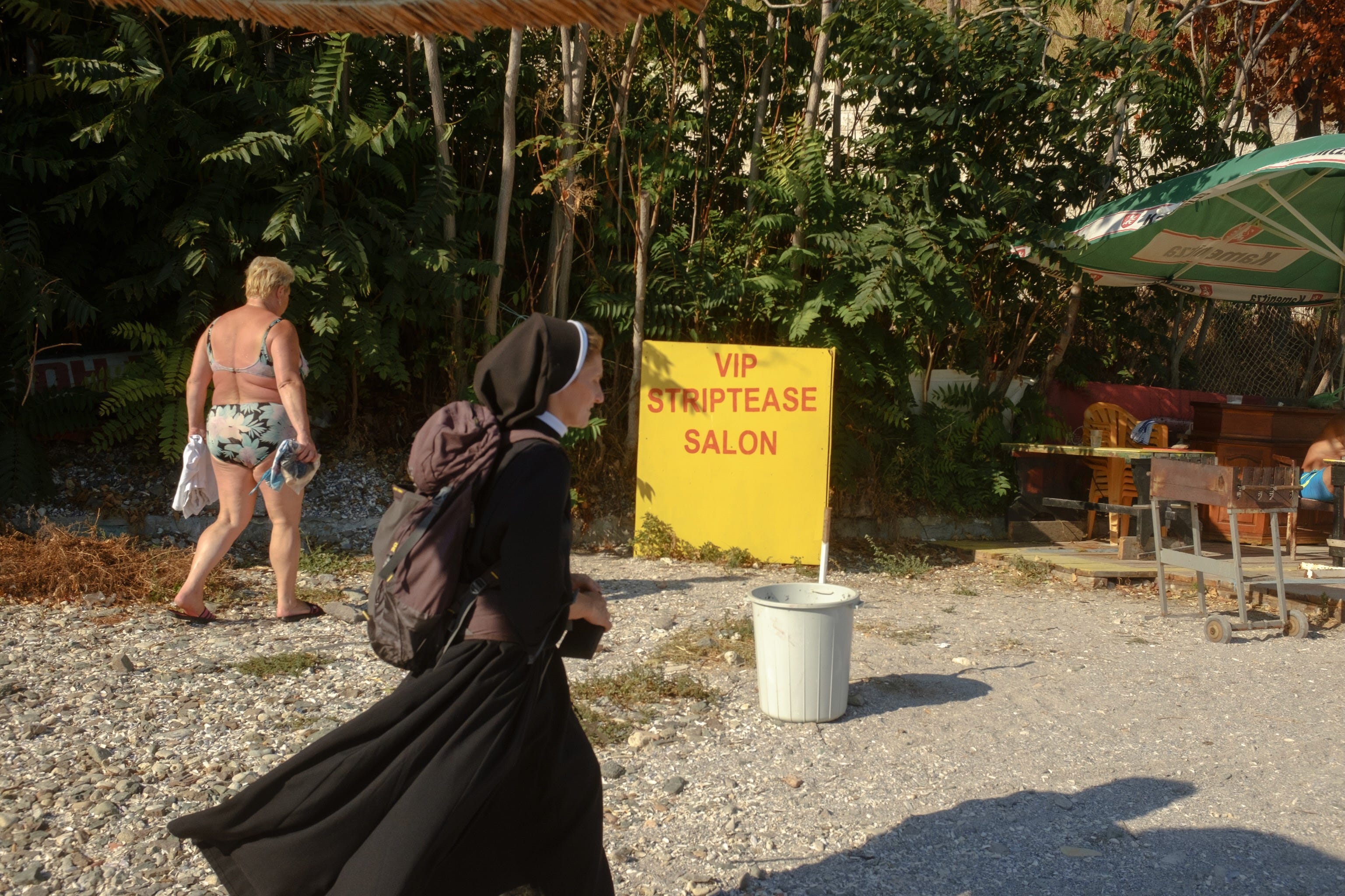 A nun and a woman in a swimsuit walk past a "VIP Striptease salon" on a beach