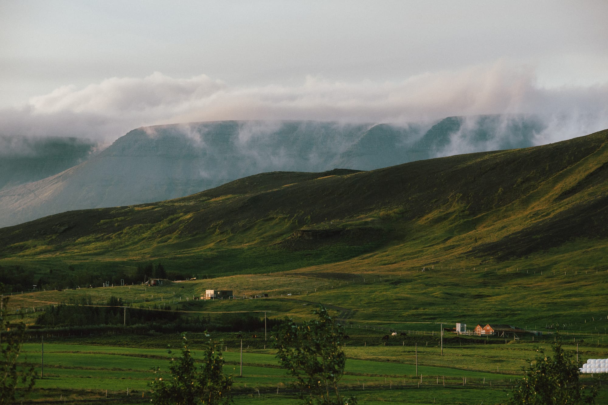 Clouds roll over a mountain in the distance. In the foregound houses seem tiny compared to the mountain ranges.