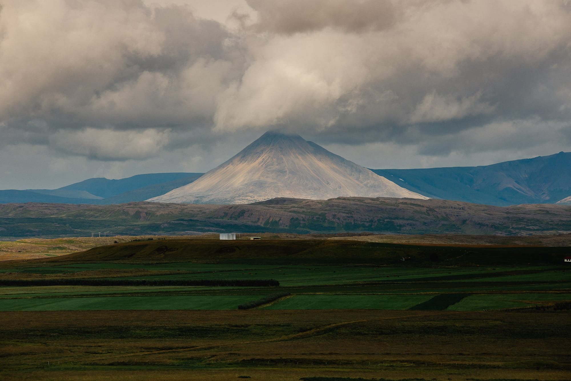 In the foreground there are rocks on a barren landscape. In the distance jagged rocky hills leading to snowcapped mountains.