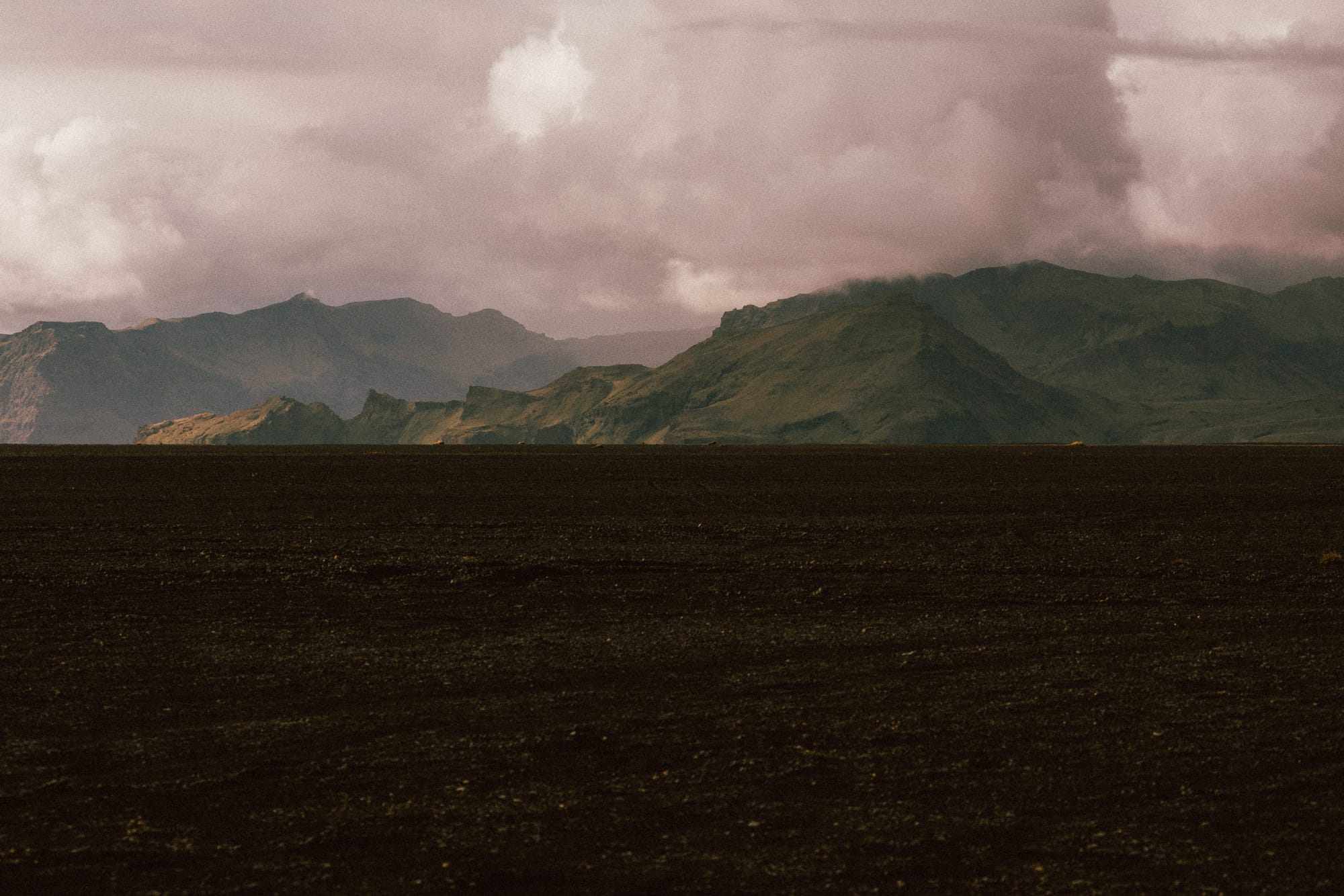 Mountains sit on the horizon of a black sandy beach.