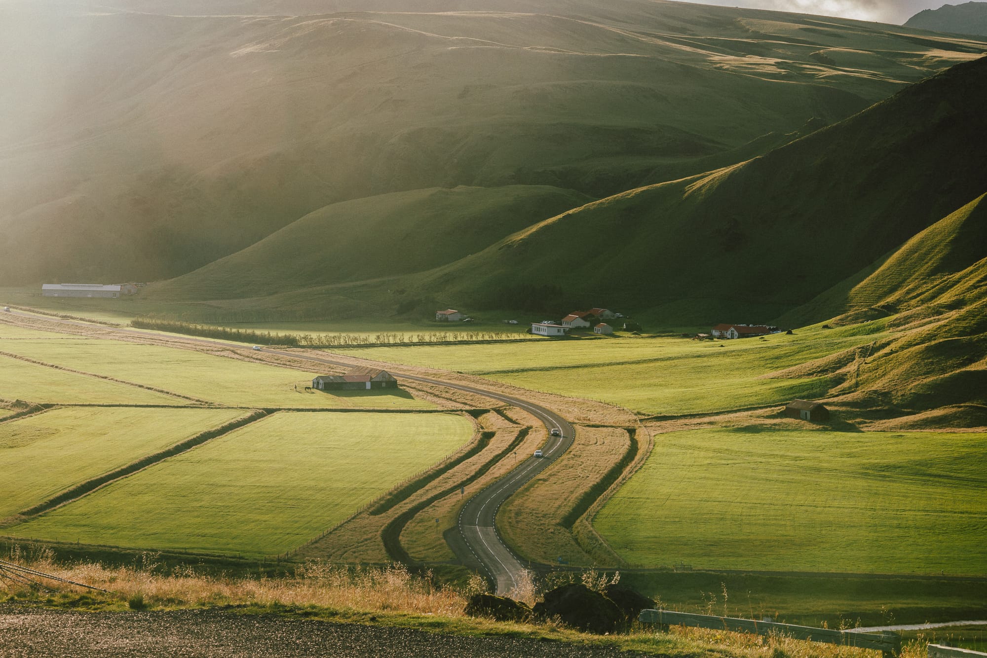 A road weaves between green fields with mountains in the background.