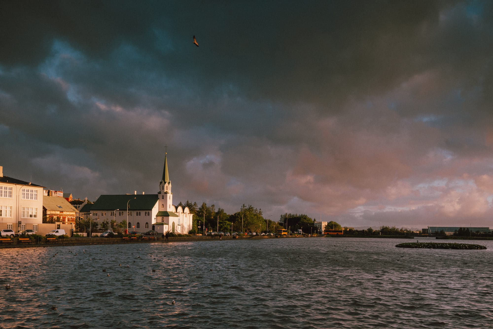 As the sun sets a church is nicely lit against a dark and stormy sky by a lake.
