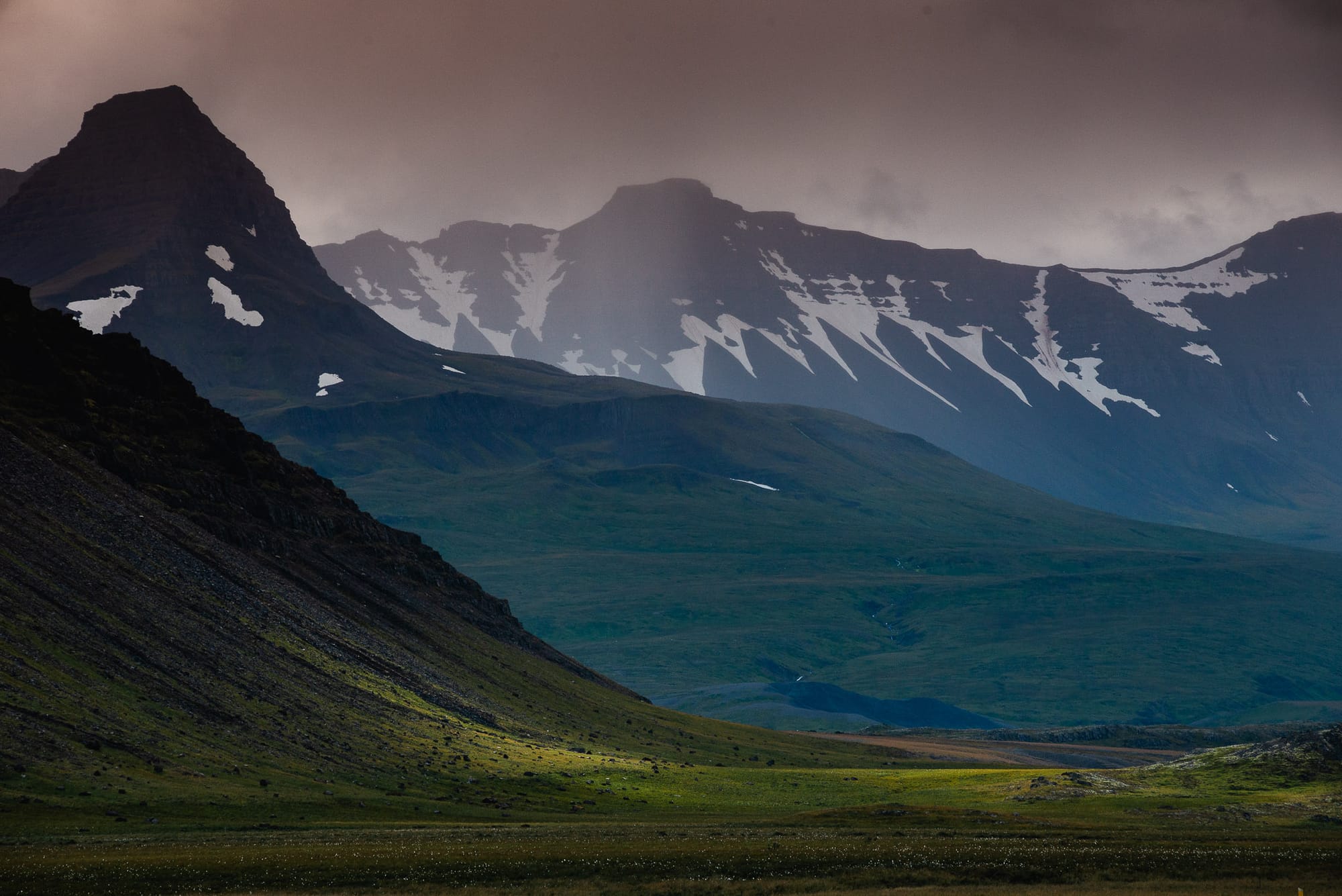 Sunlight hits fields of green grass at the bottom of a mountain range that rises up to snowcapped peaks.