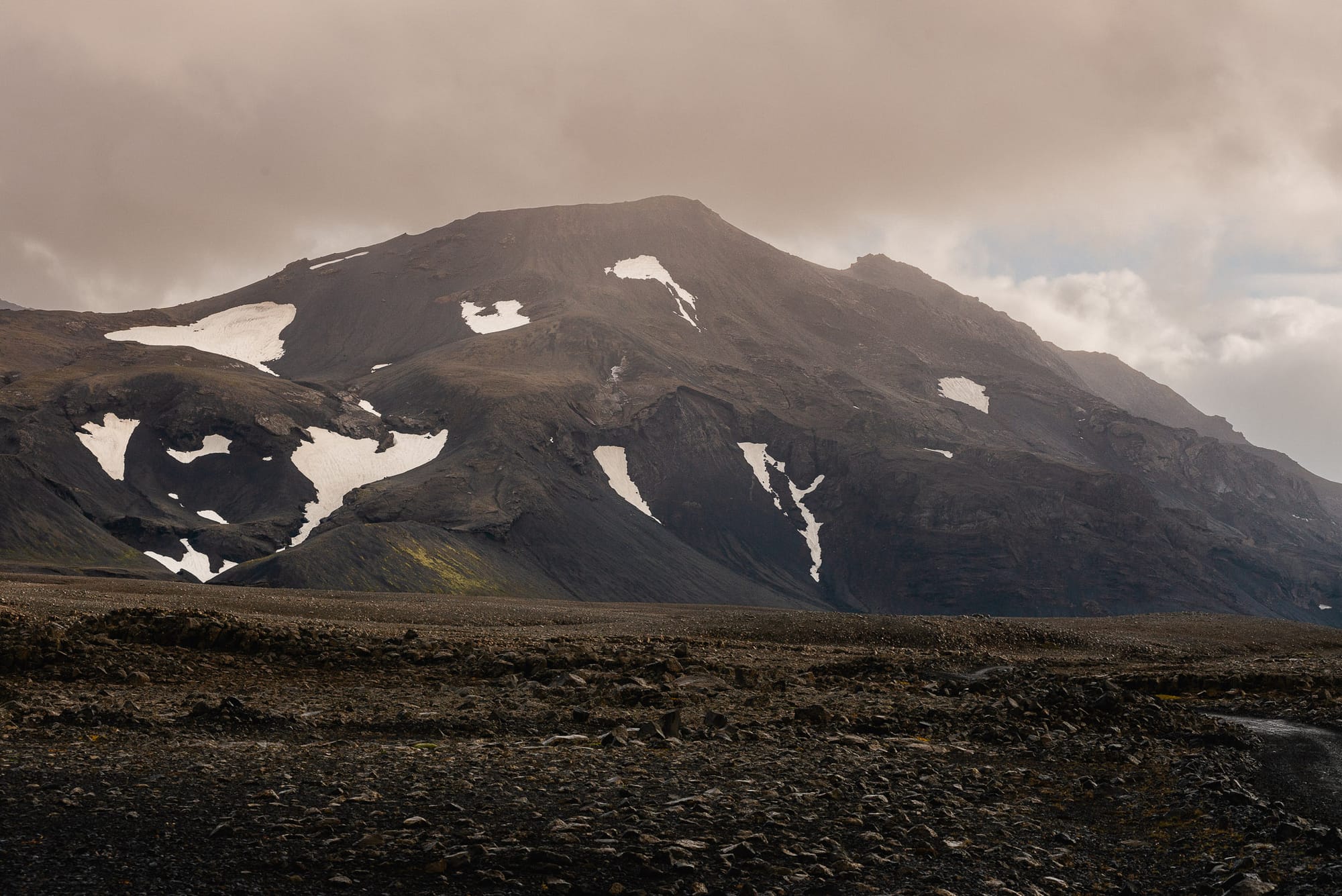 A mountain with numerous patches of snow sits on top of a volanic landscape on an overcast day.