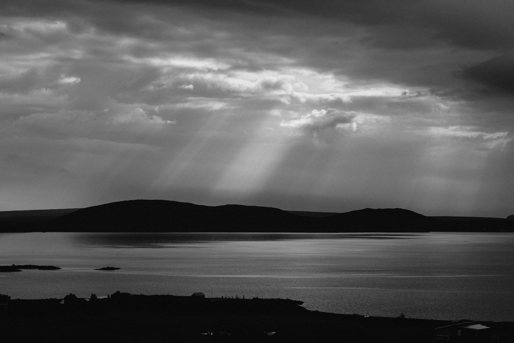 A few small islands in a lake in Iceland with mountains in the distance. As the sun sets rays of stream through the clouds.