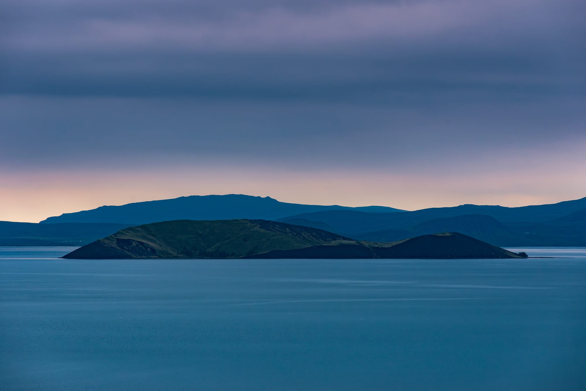 A small island in a blue lake at dusk with layers of mountains in the distance.