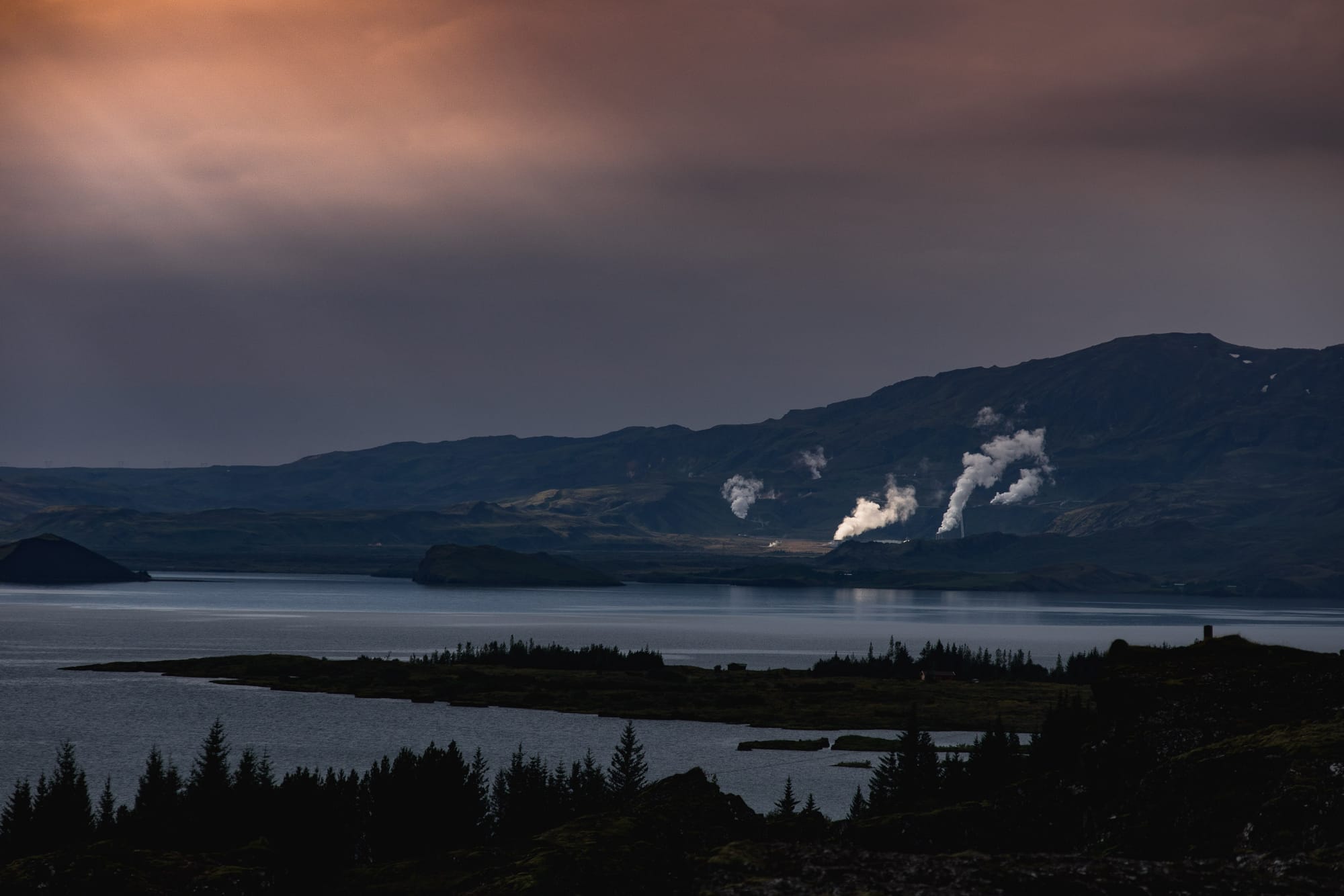 Trees line a lake. In the distance steam rises from a geothermal powerplant as streams of light break through the clouds.