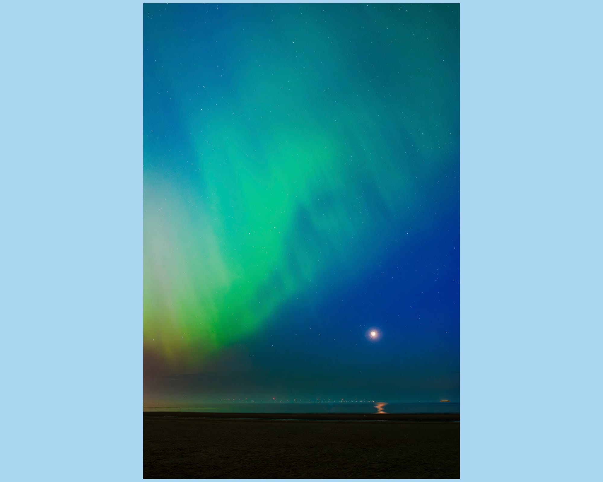 Green bands of light streak diagonally across the nights sky above a beach while the tide is out.