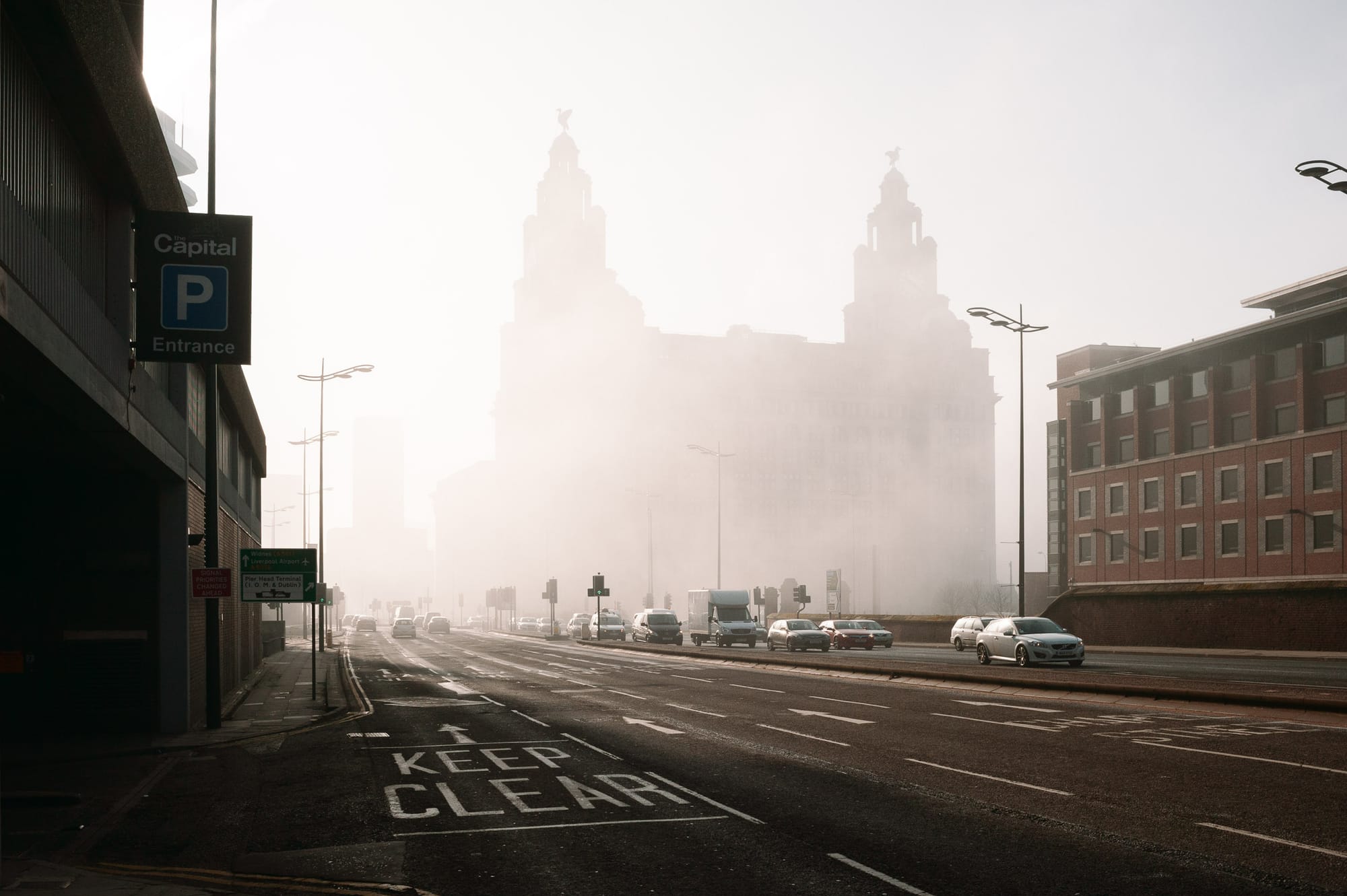 The Liver Building stands faded by early morning fog. Cars drive on either side of the road at rush hour.