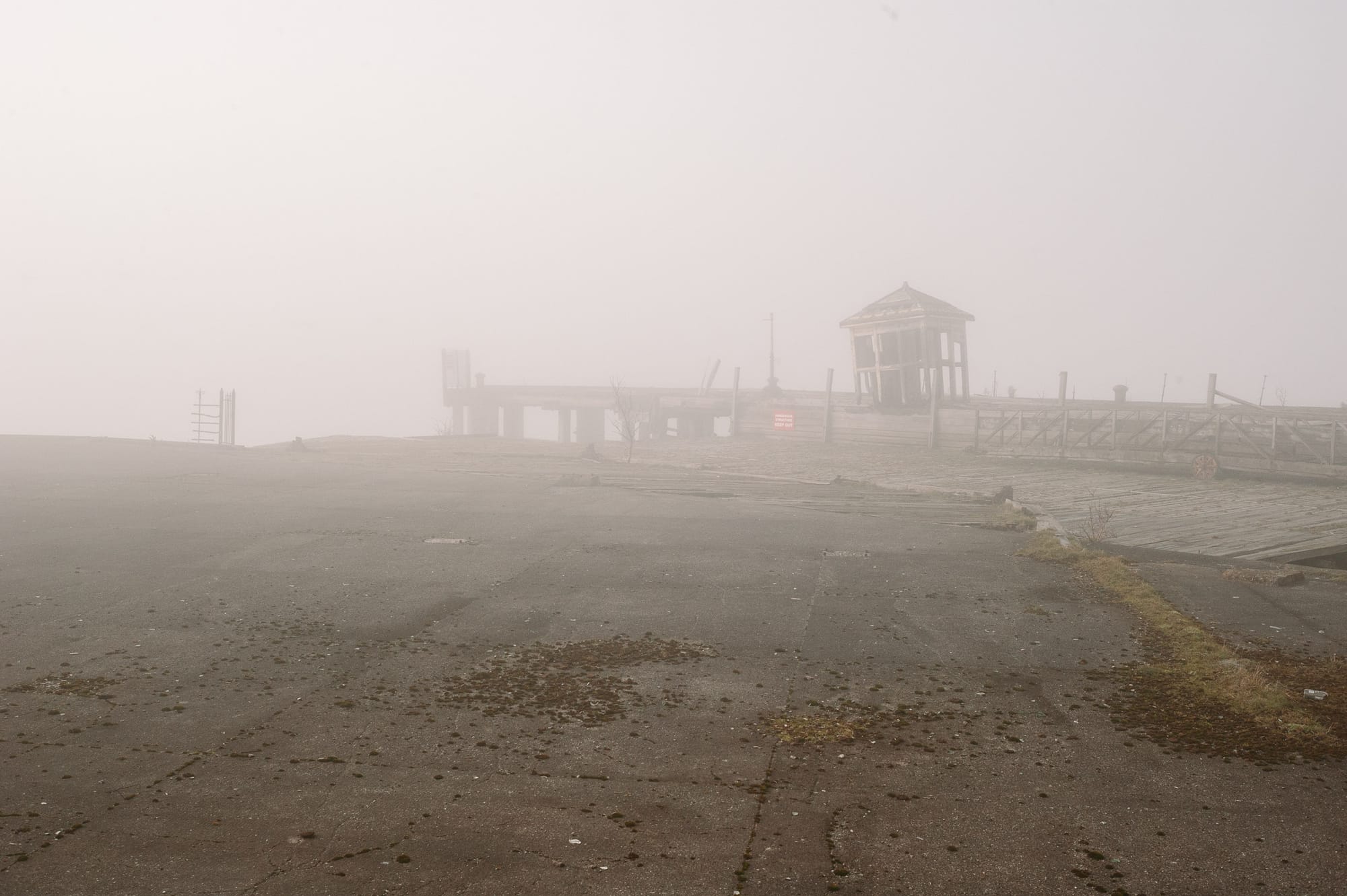An old abandoned dock disappears into fog. A small hut sits at the end of the dock with parts falling off.