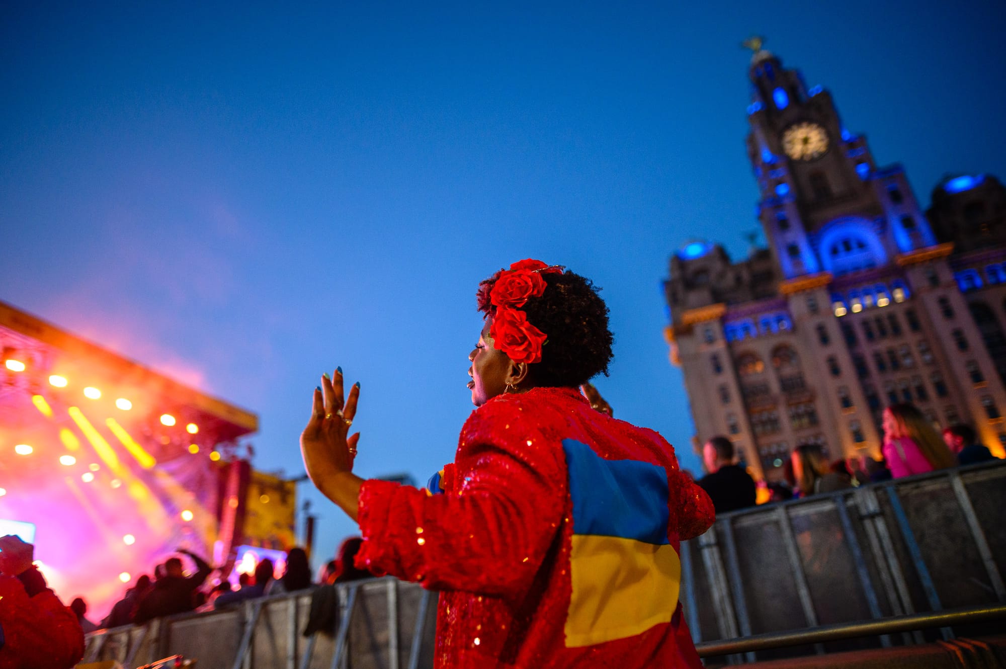 A woman dances after sunset on the Liverpool waterfront. She is wearing a red sequin jacket with the Ukraine flag on.