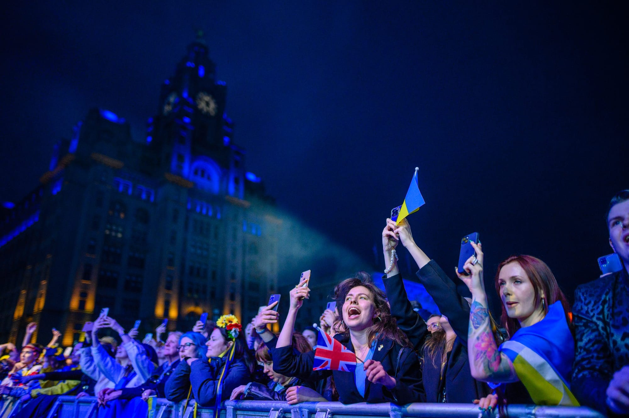 Dusk on the Liverpool waterfront. The Liver Building is lit up yellow and blue for Ukraine. There is a large group of people watching a live music event. One person is shouting for joy and waving a UK flag. Around them are people holding Ukraine flags and enjoying the night.