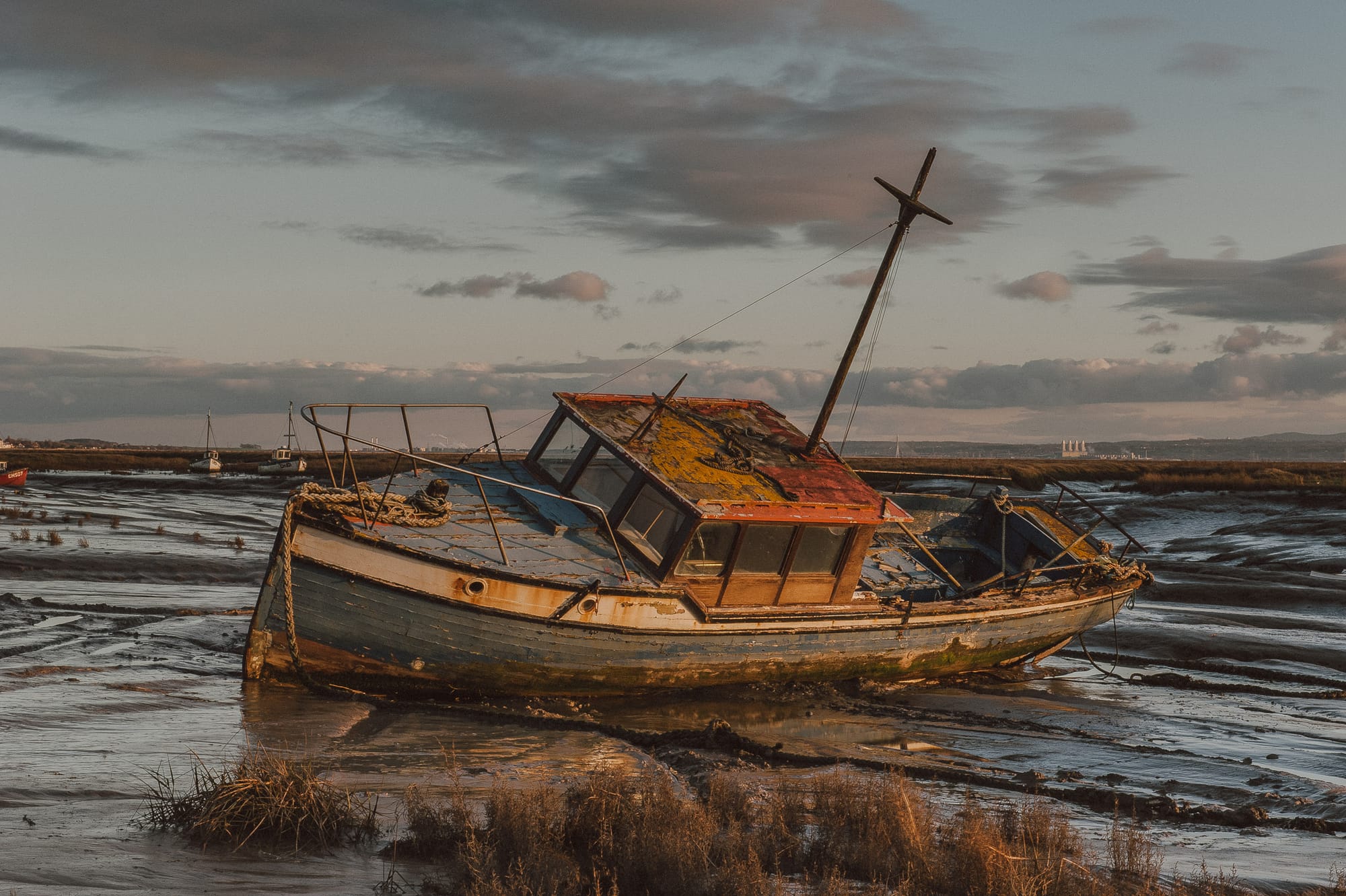 A worn out boat rests on a muddy riverbed at sunset. There are lots of bits of paint flying off and signs of rust.