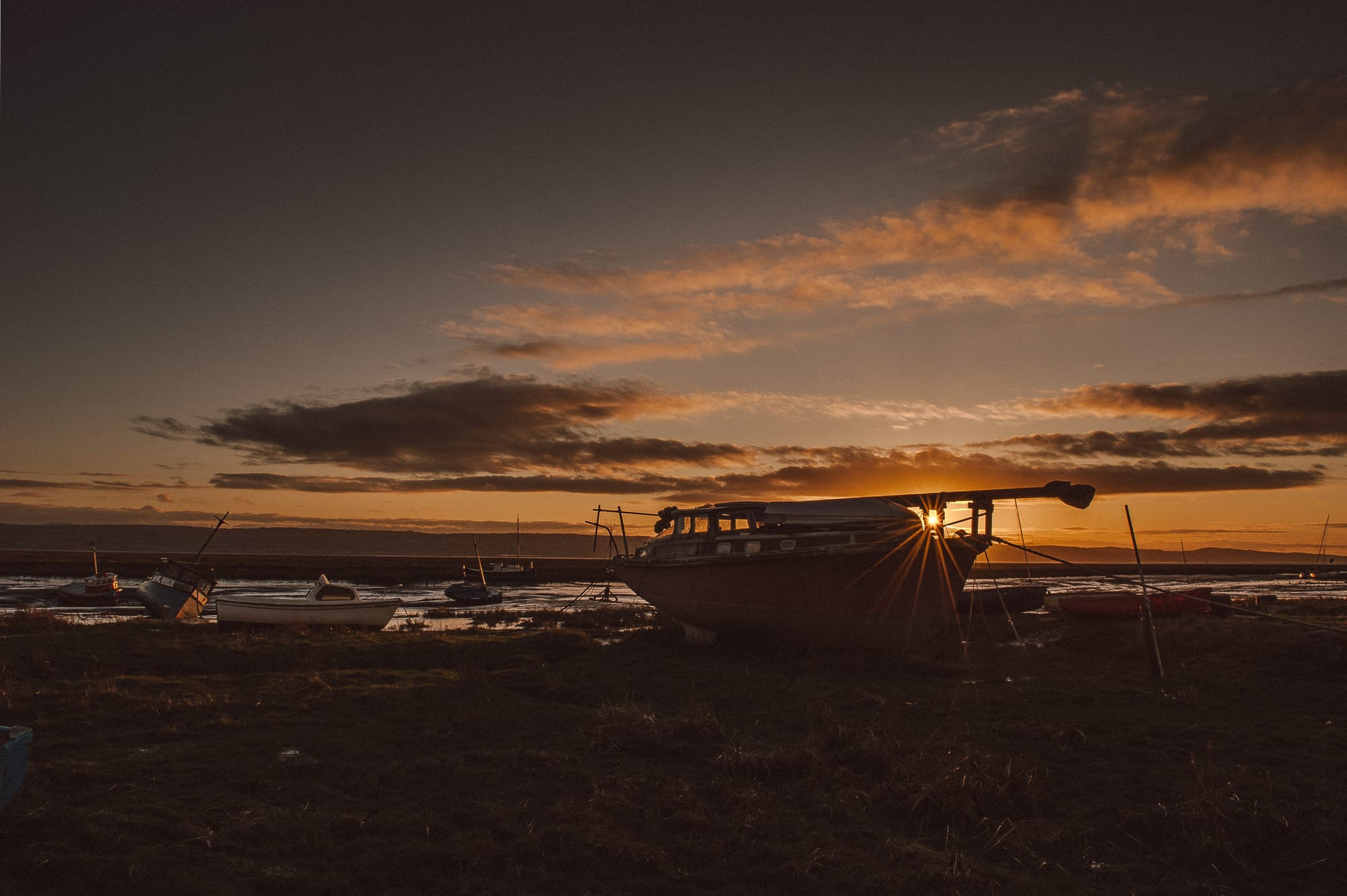 An orange sky as the sun sets behind boats resting on a grassy riverbed.