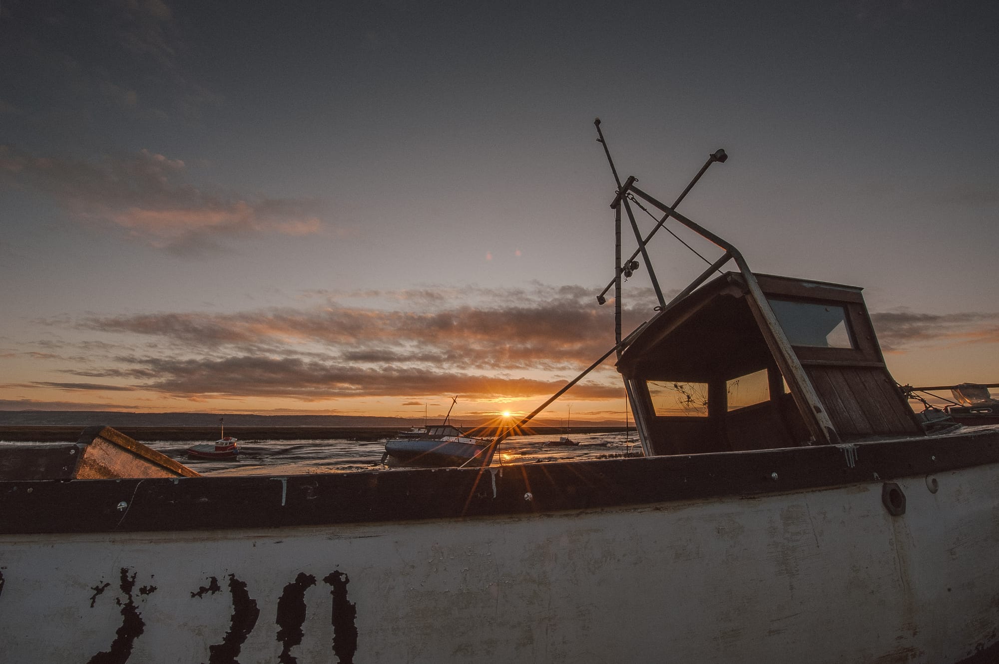 The sun sets just above an old boat resting on a dry riverbed. There are other boats in the distance.