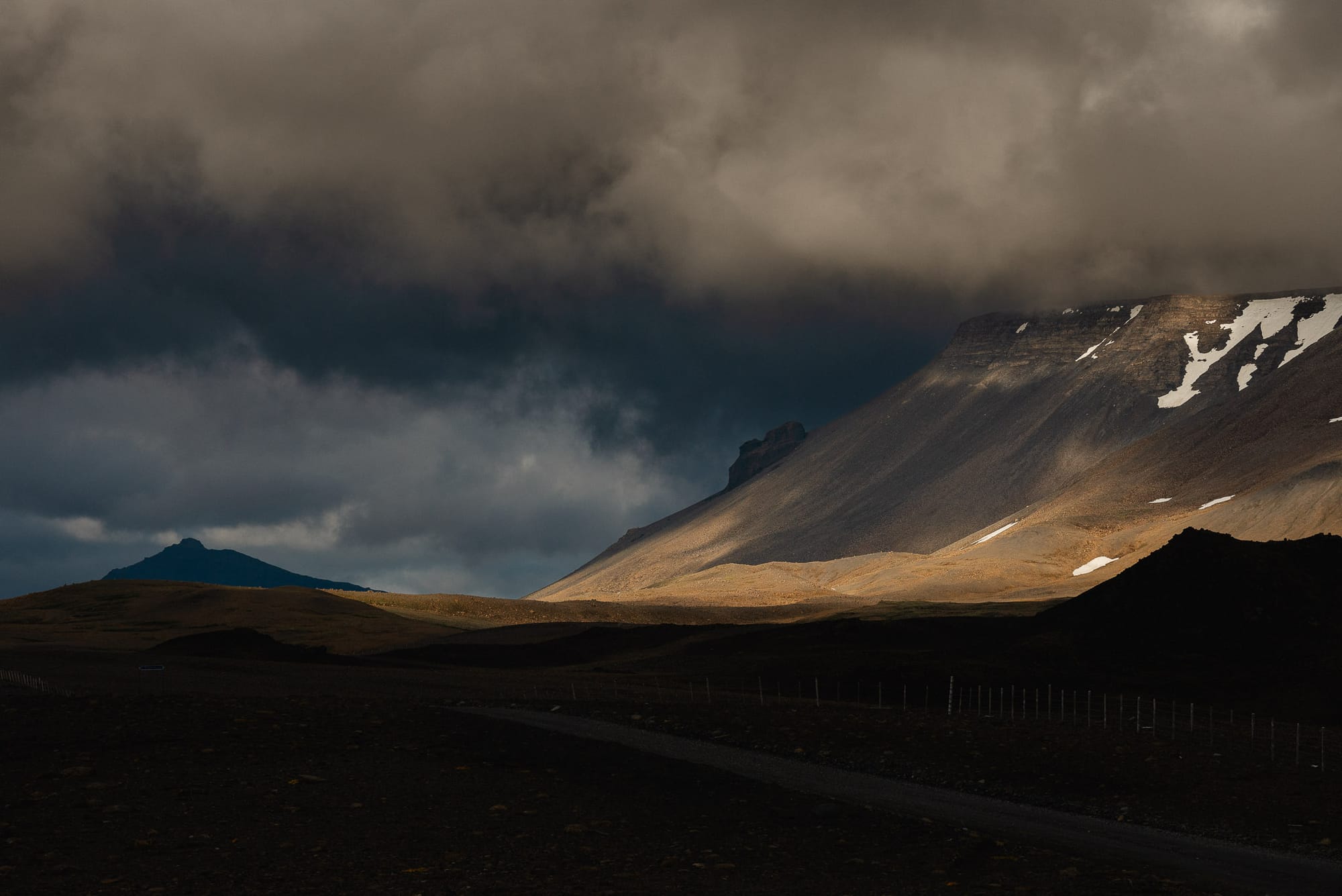 Sunlight lights up the side of a flat-top mountain. There are patches of snow on the top.
