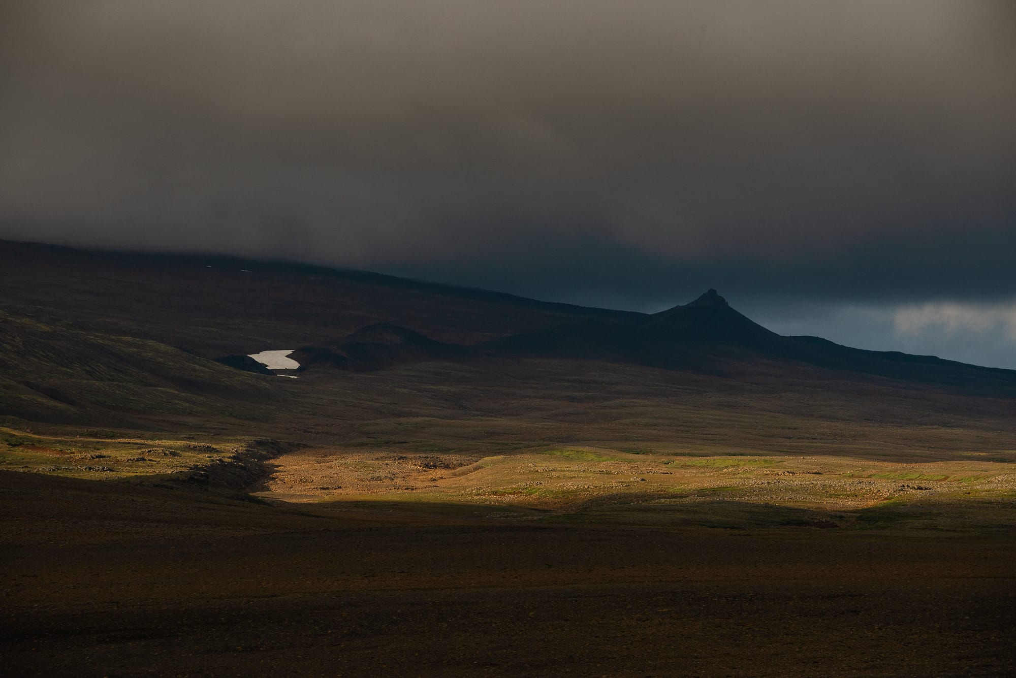 A strip of light passes over a rocky brown landscape. There is a small patch of snow and a pointy mountain in the distance.