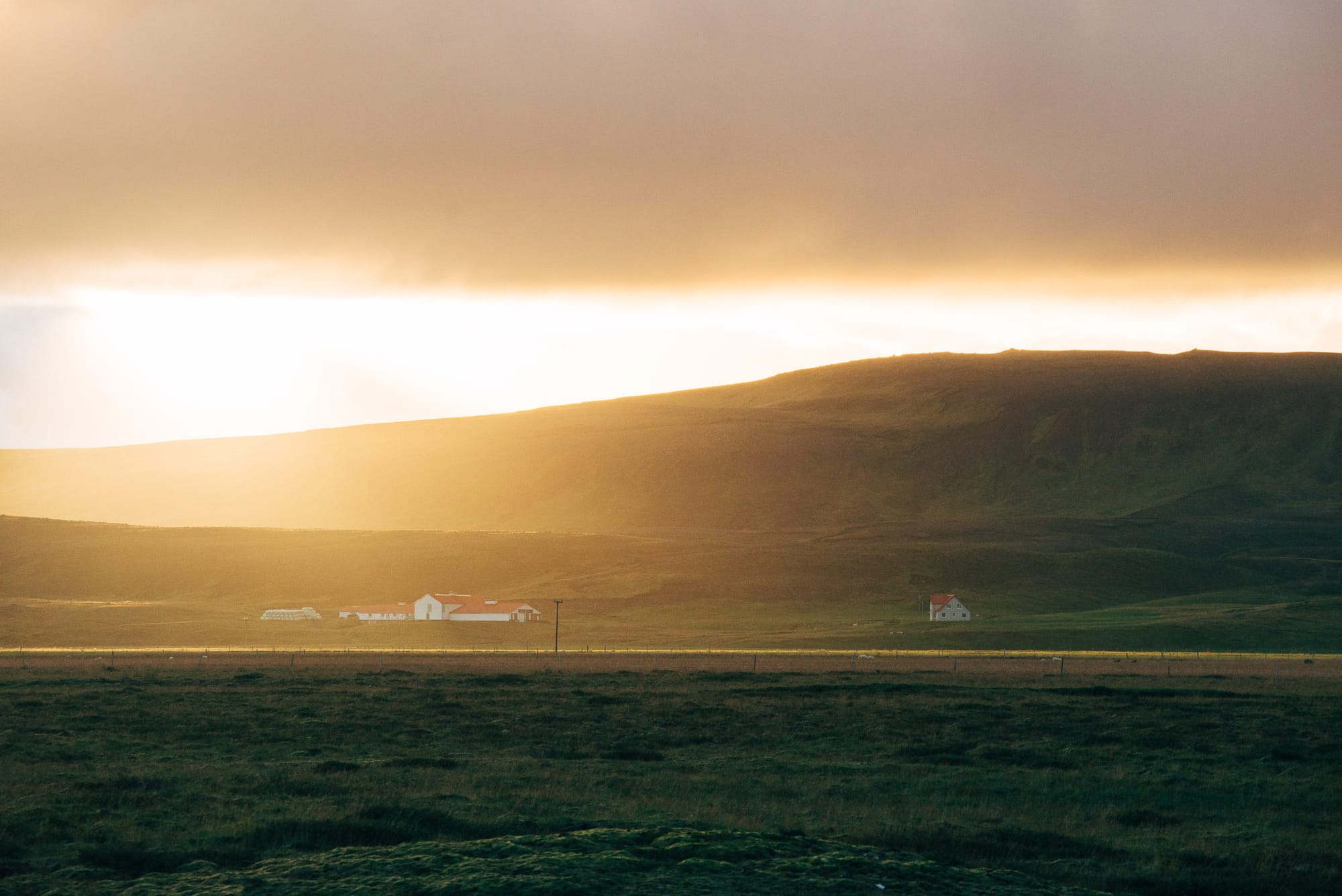 As the sun sets behind large hills, a small farm is backlit and glowing in the evening light.