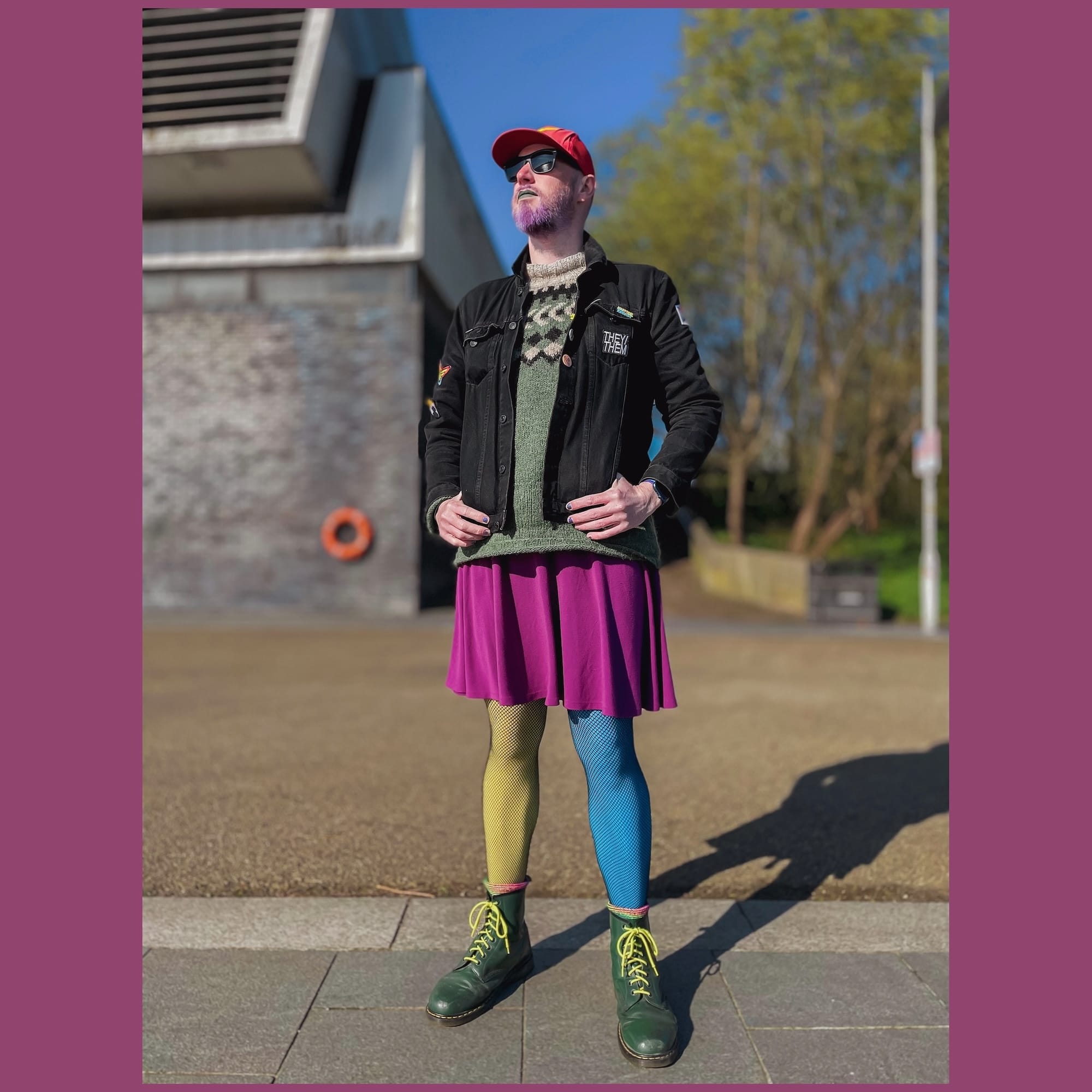 A non-binary person in green DM boots with yellow laces, Ukraine coloured fishnet tights and a pink skirt. They have a green knitted jumper on, black denim jacket with a "They/them" patch. They have a purple beard with green lipstick on, shades and a red baseball cap.