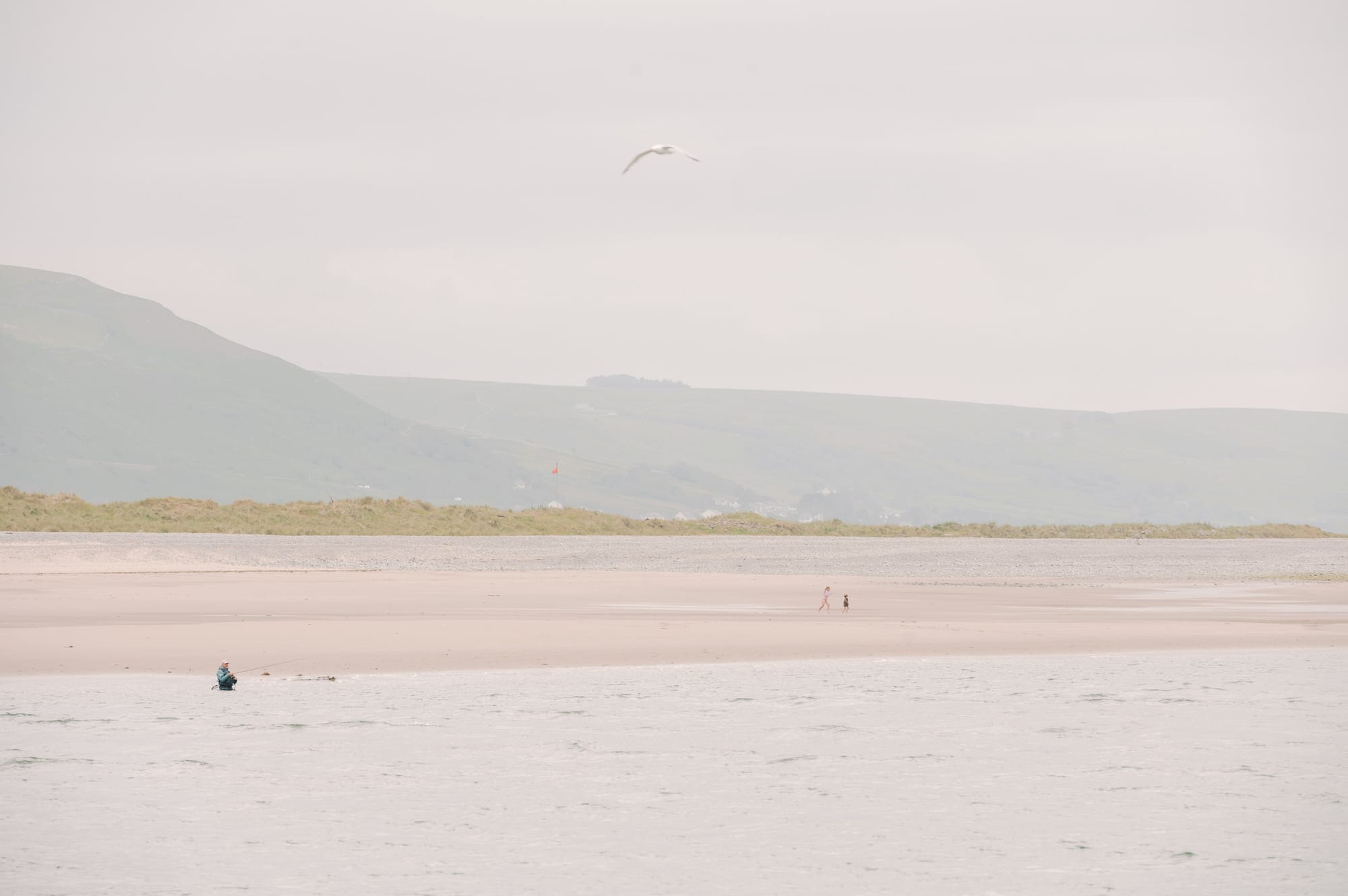 A man stands waist height fishing in an estuary while kids run around on the beach in the distance.
