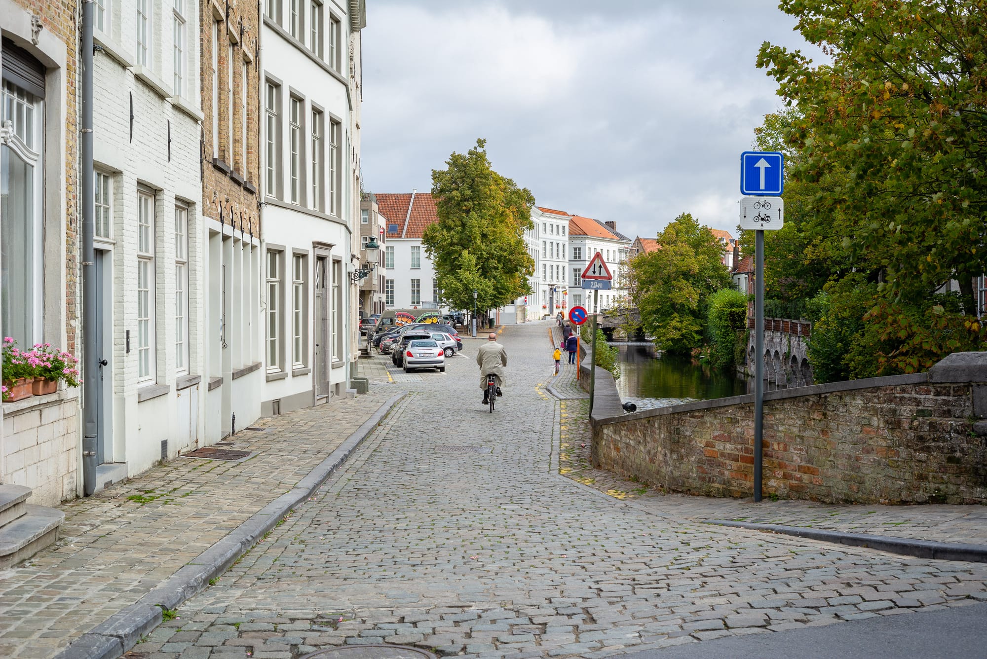A man cycles away from the camera down a lane in Bruges. On the right side is a canal and on the left a series of nice looking houses. There are lots of trees.