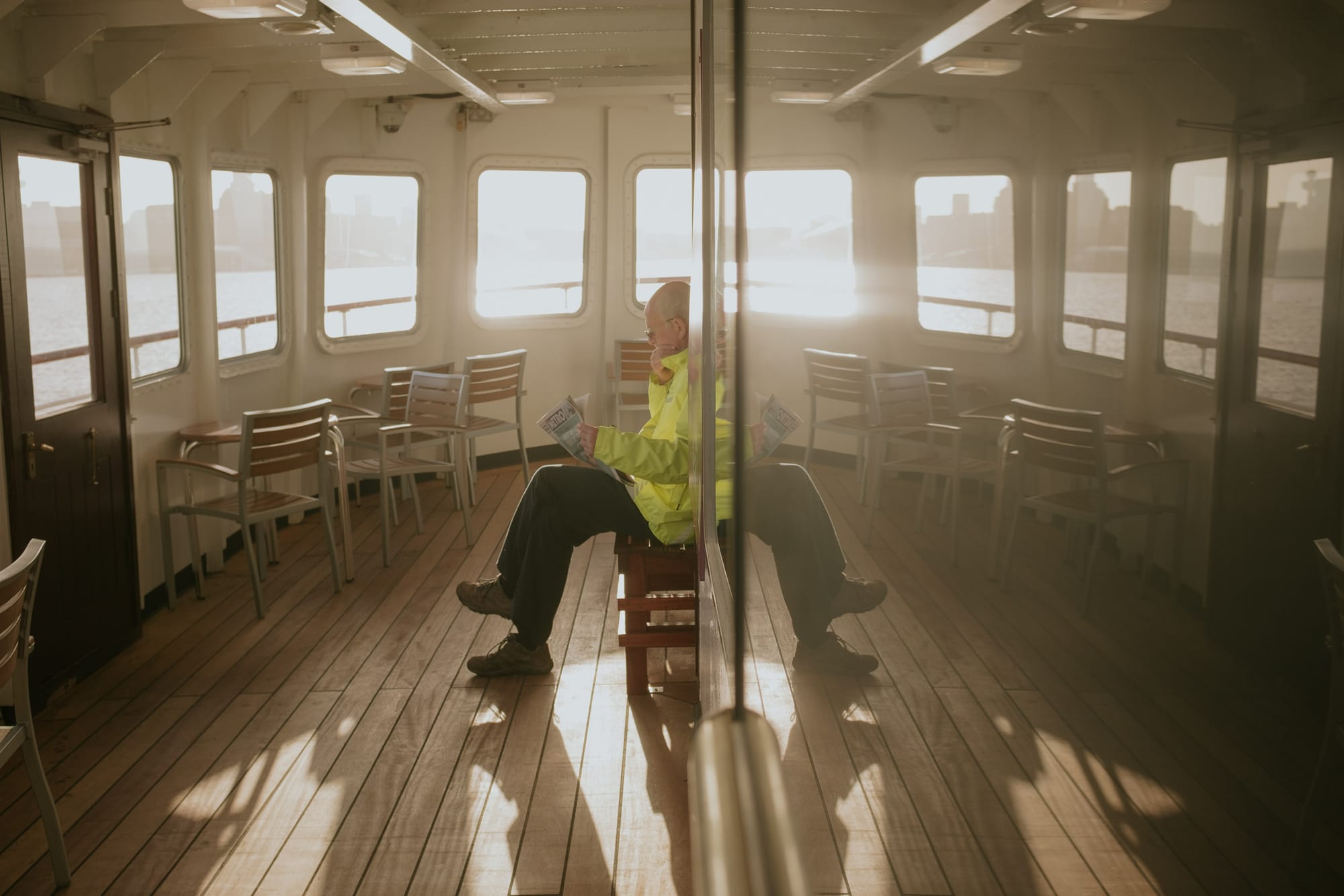 A man sits reading the morning newspaper as the Mersey Ferry sails over to Liverpool. The camera is positioned next to a reflective window to reflect the left hand side over to the right. It seems like a set of legs come out the back of the man.