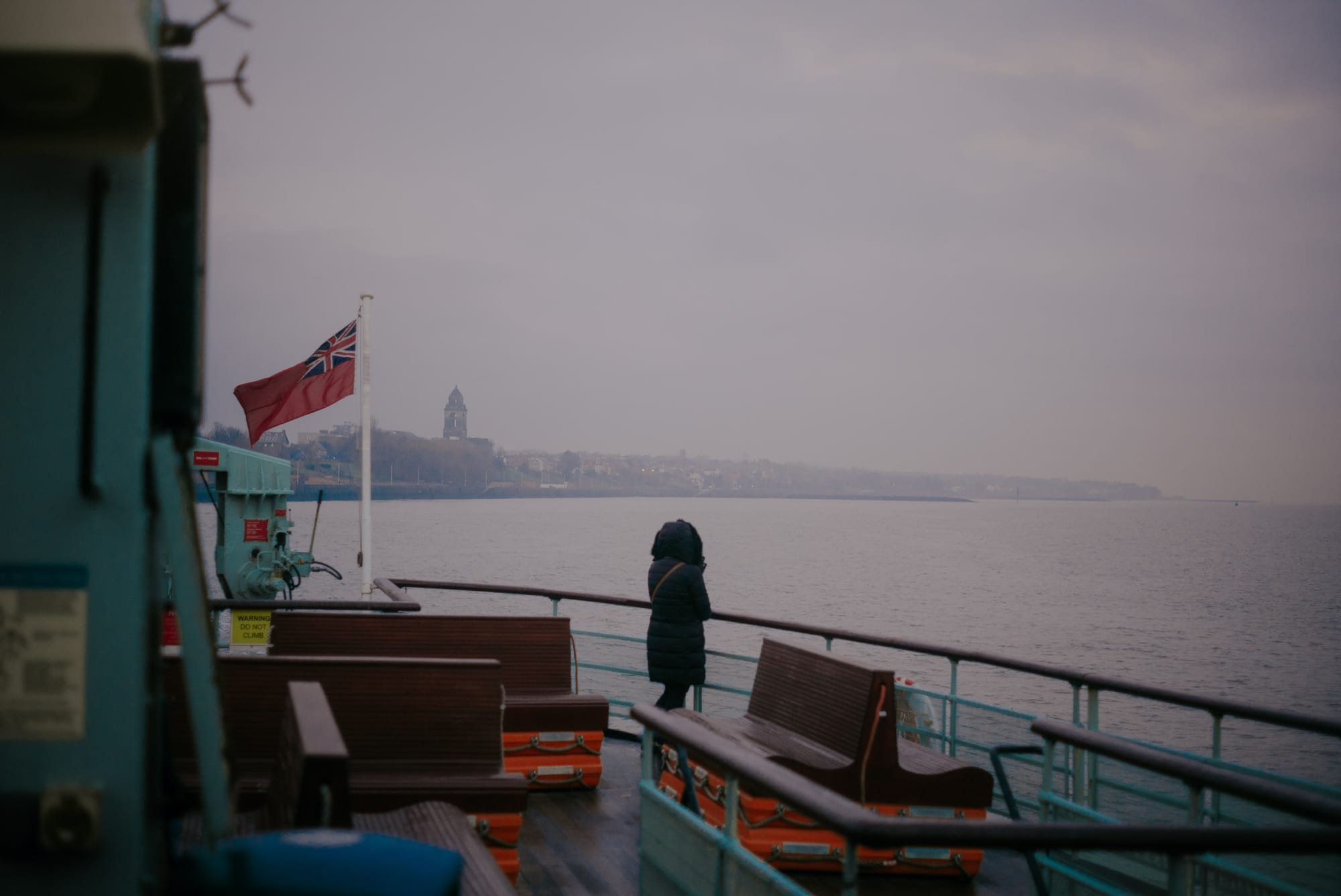 A person in a Parka jacket stands at the end of the ferry looking out over the River Mersey on a miserable day. The ferry appears quite empty.
