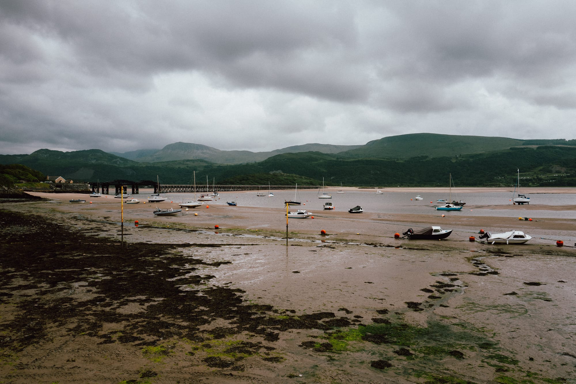 Lots of little boats rest on a river bed after the tide has gone out. Above them is a moody sky with layers of mountains fading into the distance.