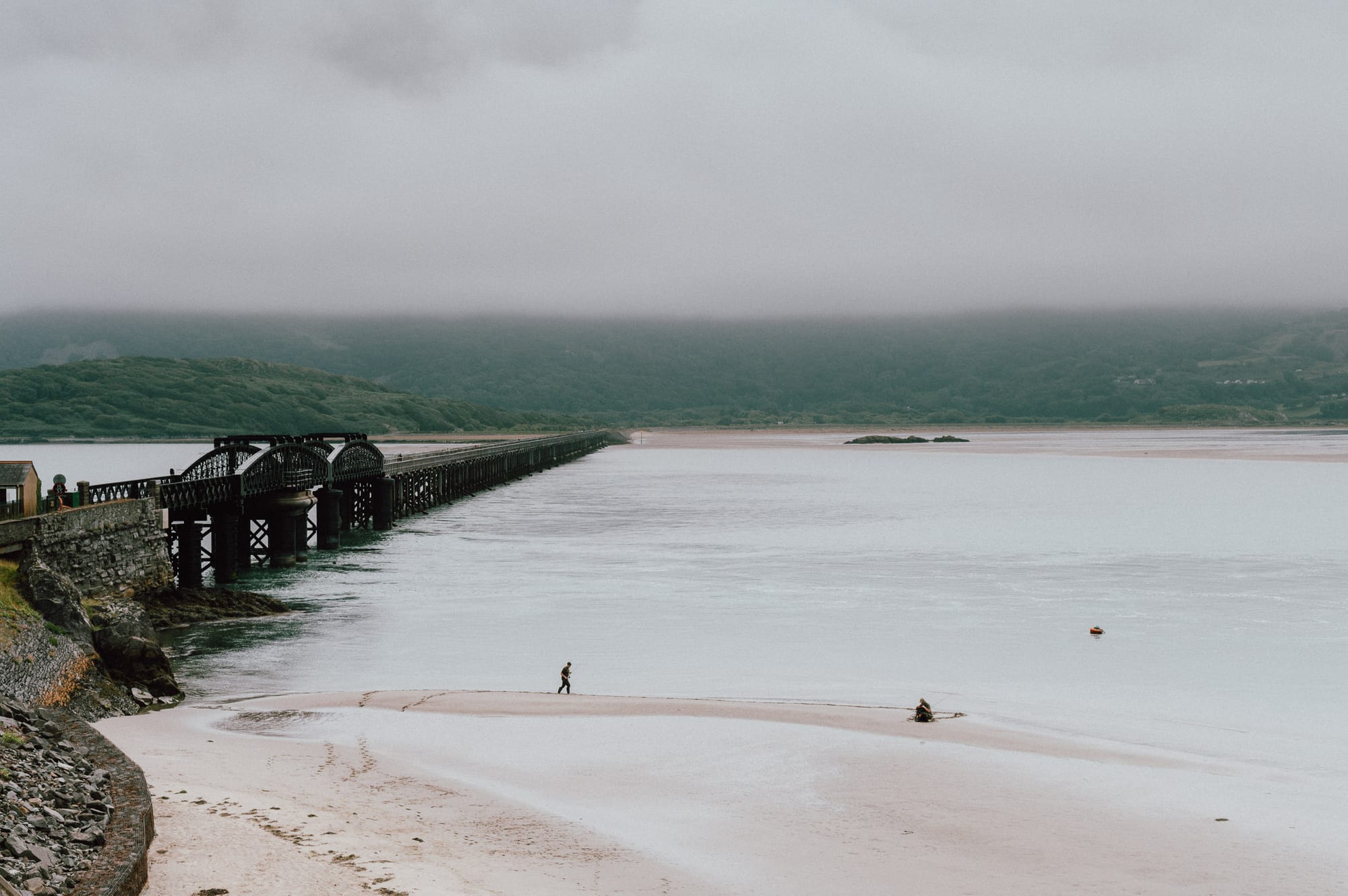 A person walks along the water’s edge towards their friend. To the left is an old railway bridge / footpath bridge. Across the water are mountains shrouded in low level cloud.