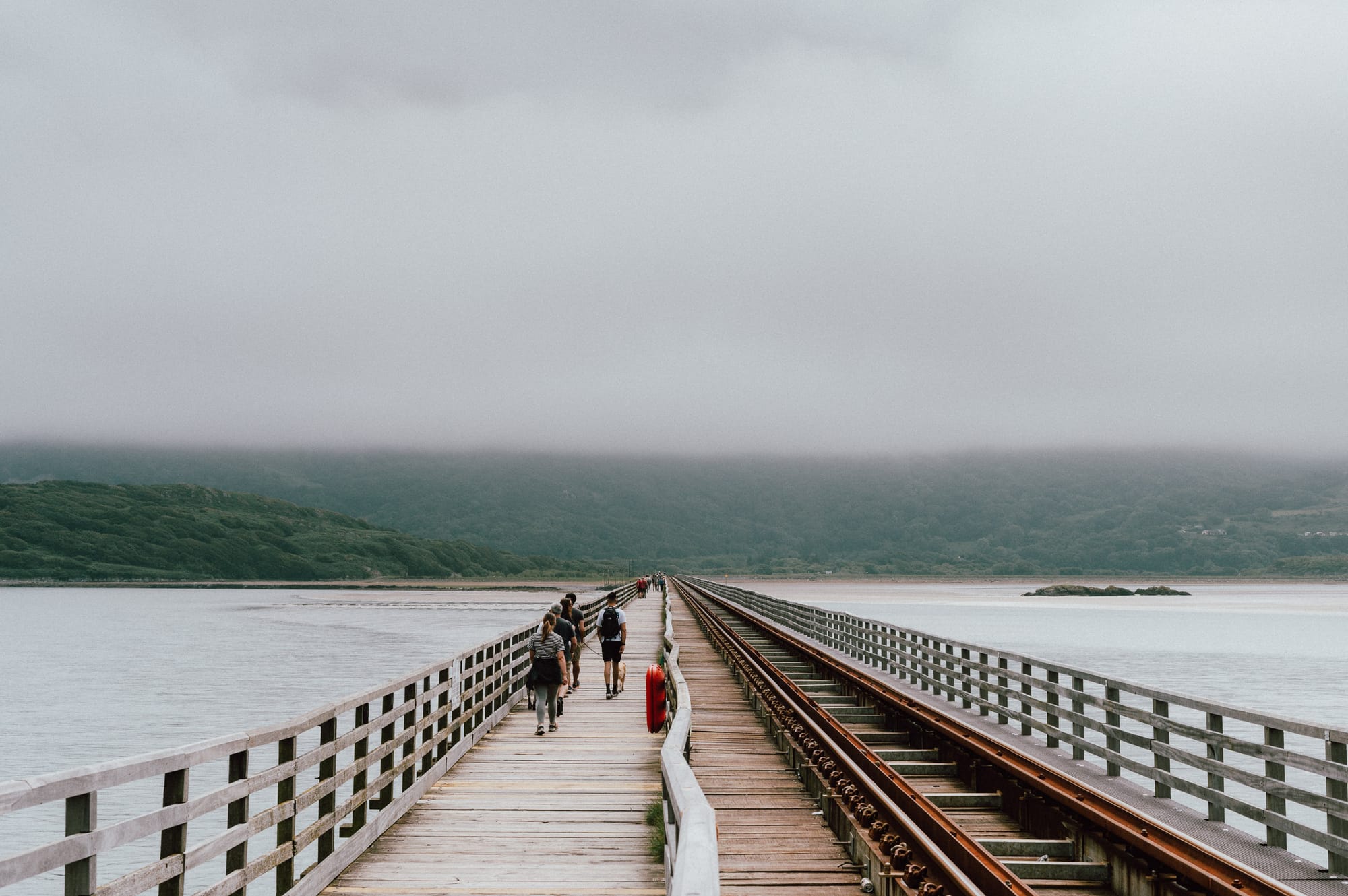 People walk across an old wooden footpath / train bridge that stretches over Barmouth estuary. In the distance clouds cover the tops of mountains.
