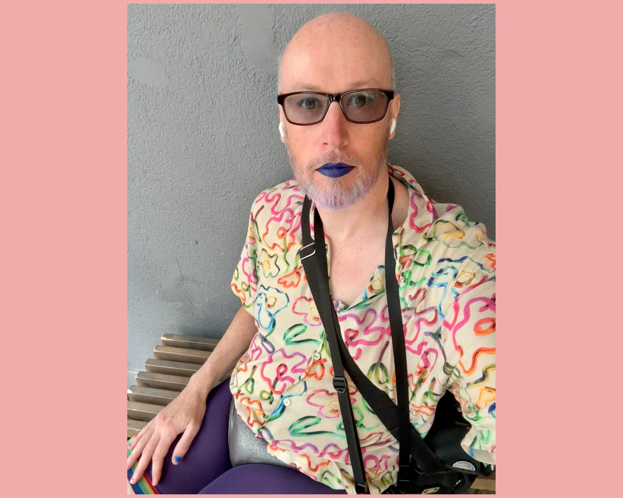 (Non-binary person wearing a shirt with rainbow coloured flower outlines on, sparkly briefs and knee length purple tights with rainbow lines at the end. They have glasses on, blue eye liner and dark blue lipstick on.)