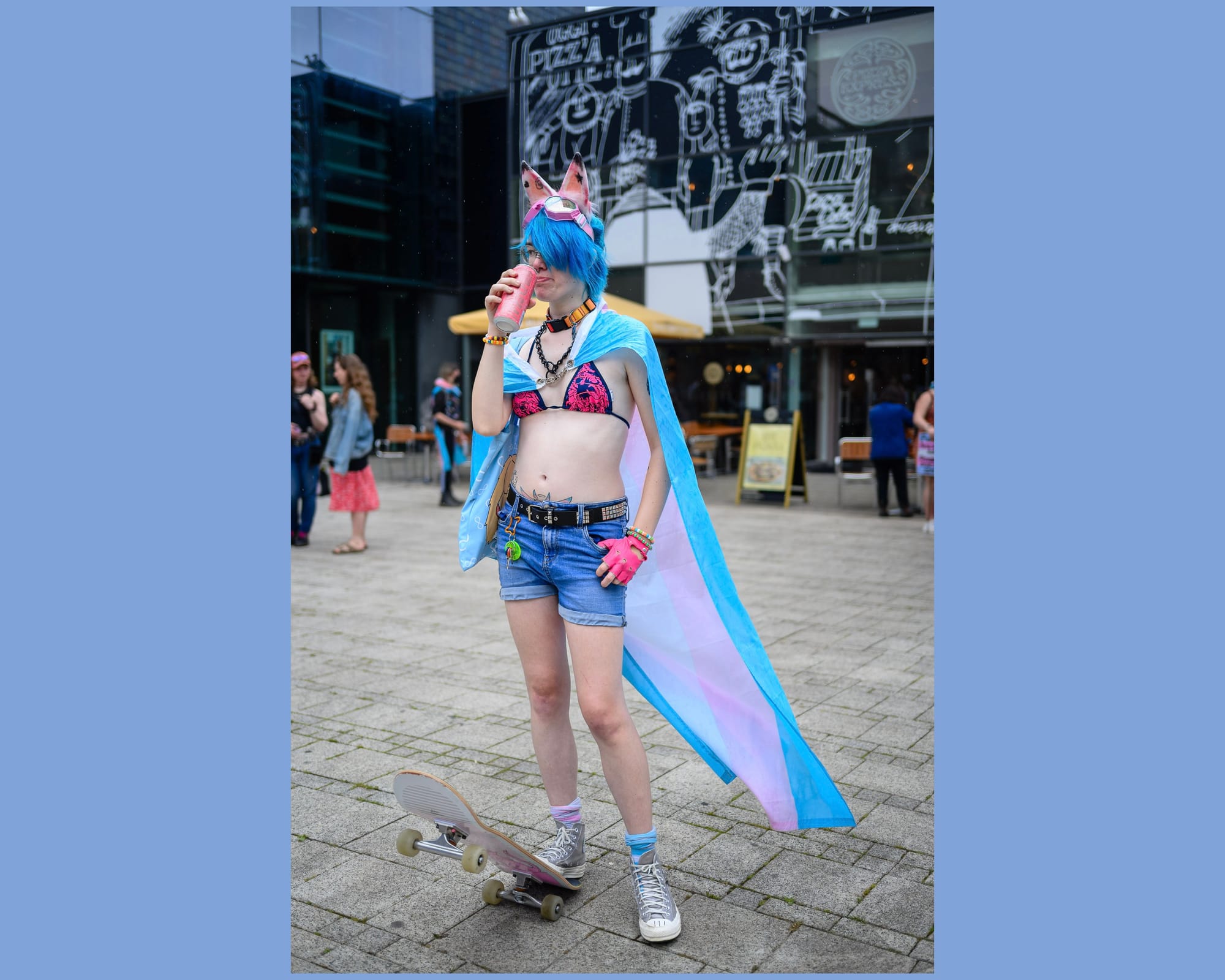 A white person casually stands wearing a Trans Pride flag as a cape with their foot on the back of a skateboard while drinking an energy drink.