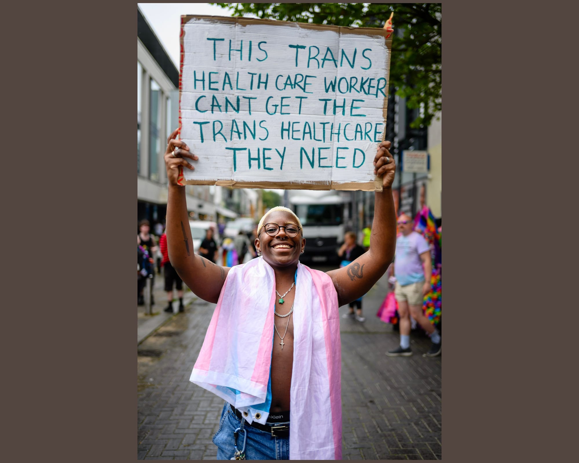 A black trans man with the trans flag draped over their shoulders holds up a sign saying “This trans health care worker can’t get the trans healthcare they need.”