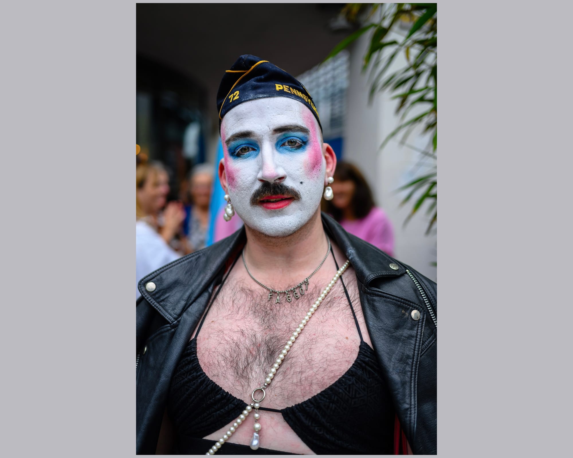 A person has their face painted white with blue eye shadow and pink tinted cheeks. They’re wearing a leather jacket which is open showing a hairy chest and knitted bra.