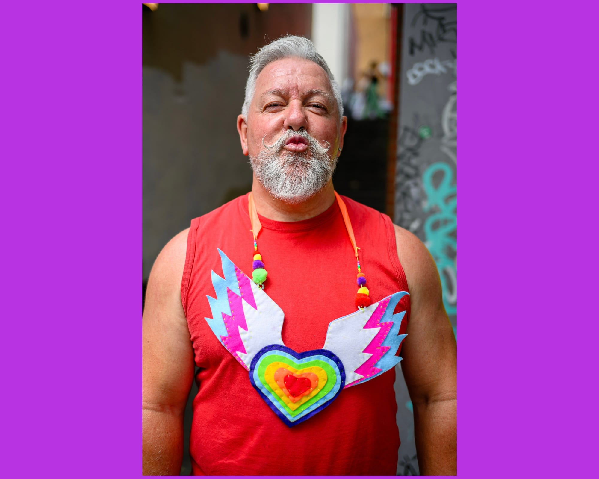 A person gives a cheeky kiss expression to the camera. They have a great beard and a large winged heart necklace.